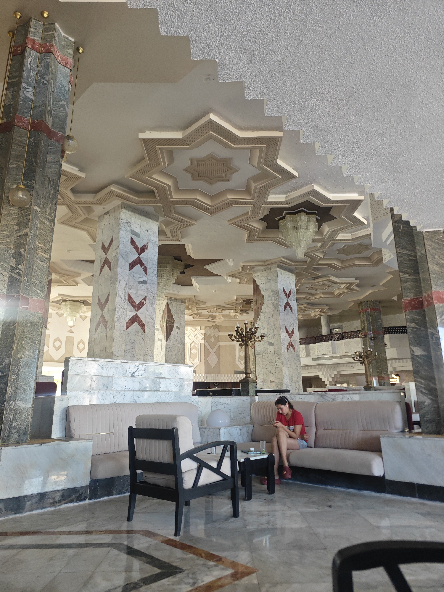In the grand lobby of a hotel in Hiboun, Tunisia, a young woman, wearing a red t-shirt and shorts, is seated on a light-colored sofa, engrossed in her mobile phone. The lobby features a striking architectural design with soaring columns clad in grey and white marble, accented with red decorative elements. The ceiling is adorned with intricate geometric patterns, reminiscent of Islamic art, and illuminated by chandeliers. Several other seating areas and decorative lamps are visible throughout the spacious lobby, which appears to be made of marble flooring. The overall ambiance suggests a luxurious and ornate setting.