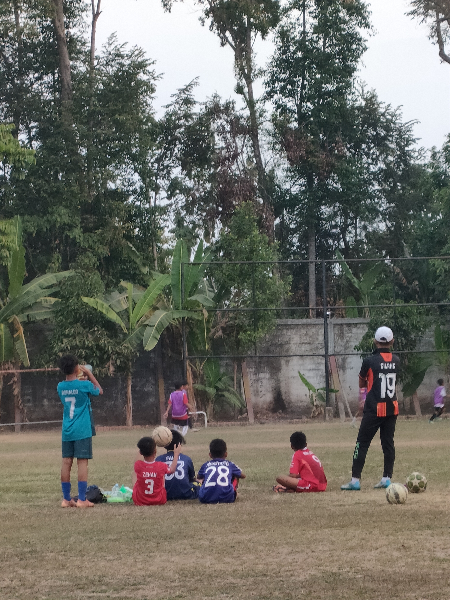 In Banguntapan, Indonesia, an outdoor soccer field is captured at an overcast dusk. A group of boys, aged approximately 7-12, are the primary subjects. Three boys are seated on the grass, wearing red, blue, and navy jerseys numbered 3, 23, and 28, respectively. Another boy in a red jersey with number 9 is also seated. One boy in a turquoise jersey with the name "RONALDO" and number 7 stands holding a ball. In the background, a boy in a purple vest is running, and another in an orange and black jersey with number 19 stands facing away from the camera. Two soccer balls are visible on the grass. The environment includes a chain-link fence, a concrete wall, and lush green trees and banana plants.