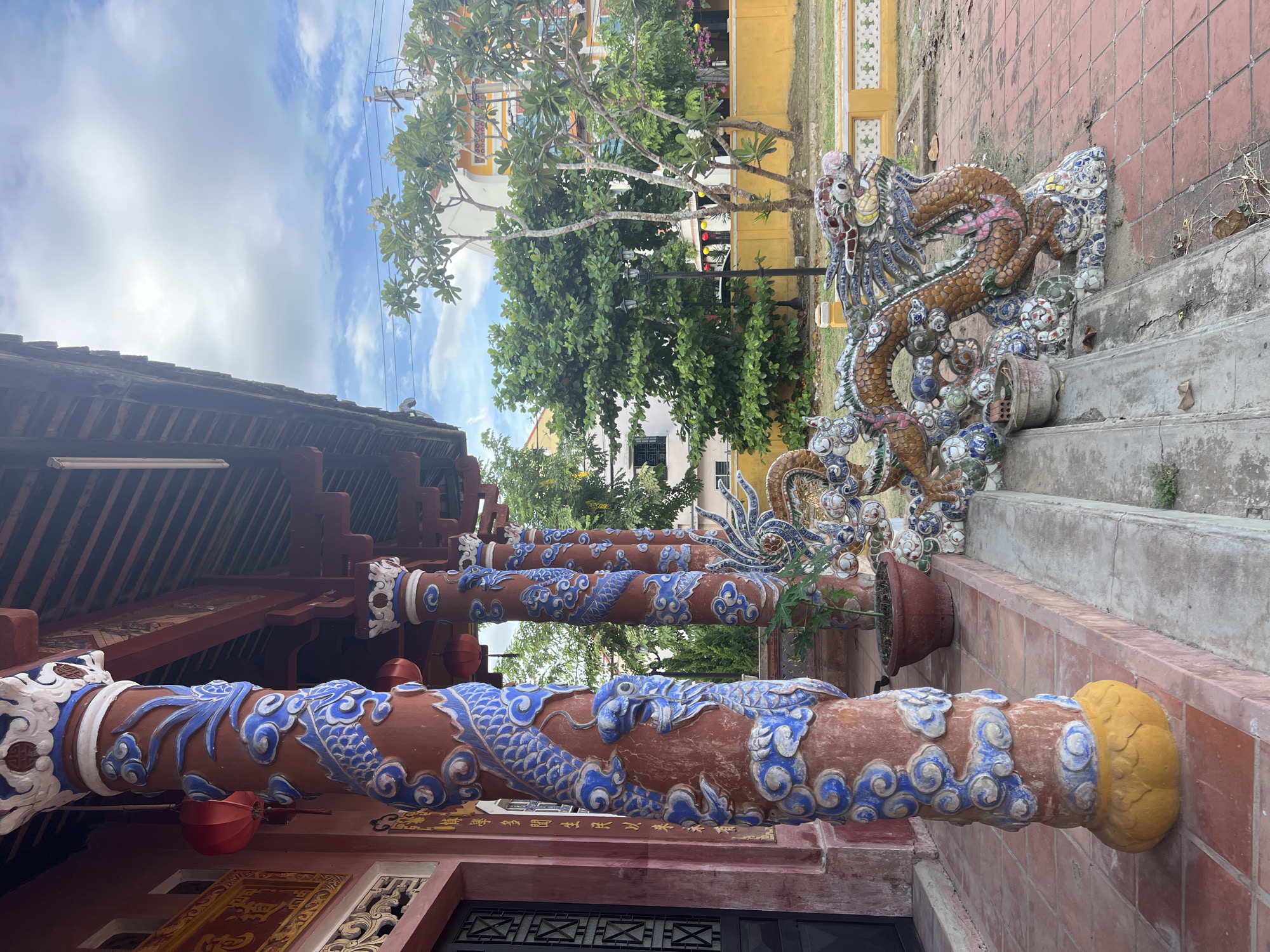 The image depicts a row of ornate, red-painted columns adorned with blue dragon motifs and floral carvings, lining the porch of a building in Hoi An, Vietnam. The columns are topped with white and blue decorative capitals. In front of the columns, there is a decorative ceramic dragon sculpture resting on a tiled step. A potted plant is visible near one of the columns. The building has a traditional wooden roof with tiled sections. Behind the building, lush green trees and a bright yellow wall are visible, with a cloudy blue sky overhead. The foreground is paved with red tiles.