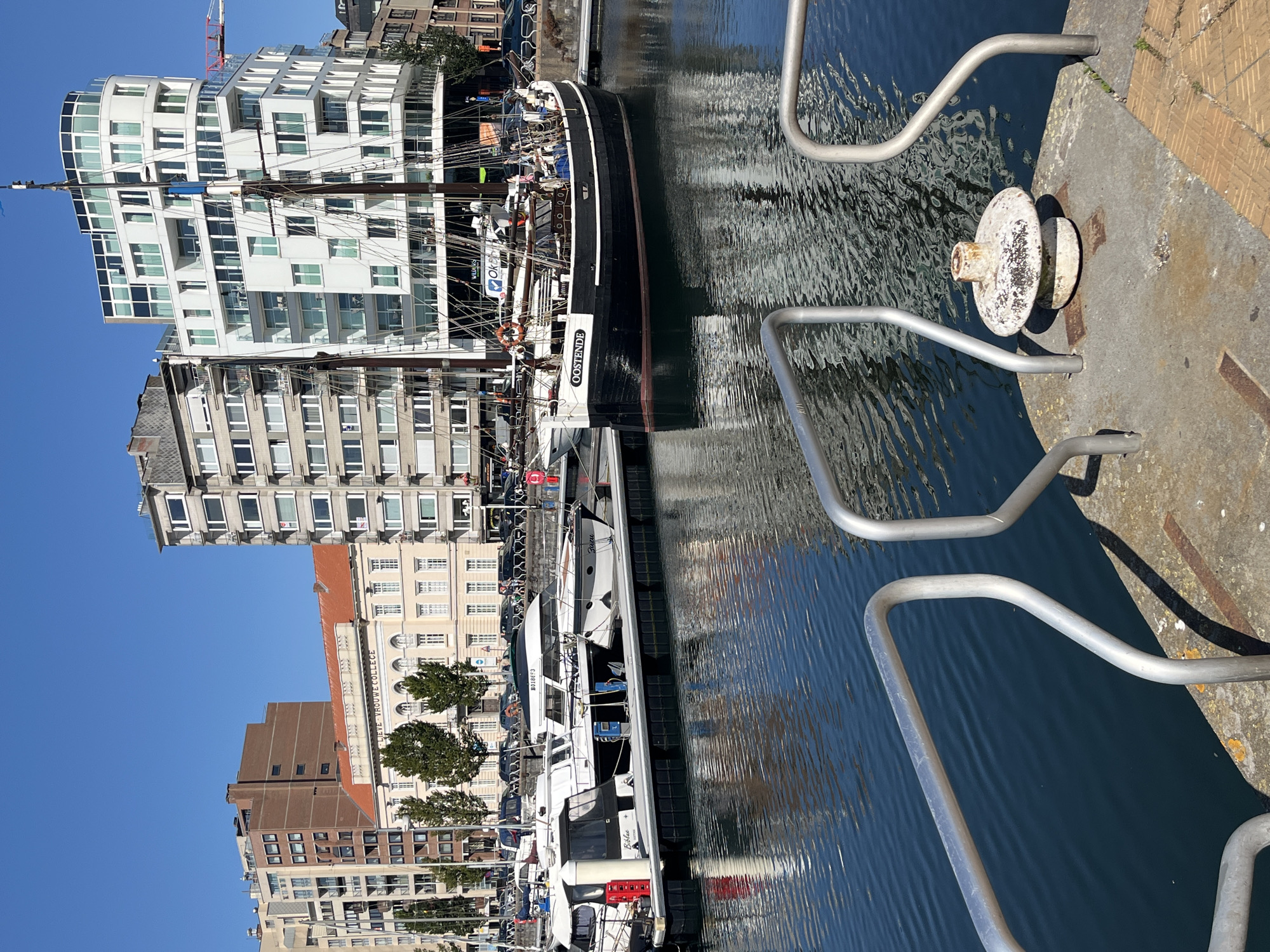 In Ostend, Belgium, on a clear, sunny day, a harbor scene unfolds with a prominent black sailing ship named "OOSTENDE" docked in the foreground. The ship, with its tall masts and rigging, is positioned alongside a modern, white, multi-story building with numerous windows. To the left of the sailing ship, several smaller white boats are also moored. The water in the harbor is a deep blue, reflecting the surrounding buildings and the sky. Along the waterfront, a row of buildings, including an older, cream-colored building with a red-tiled roof, can be seen. The background features a clear blue sky and other urban structures. The image captures a lively waterfront area.