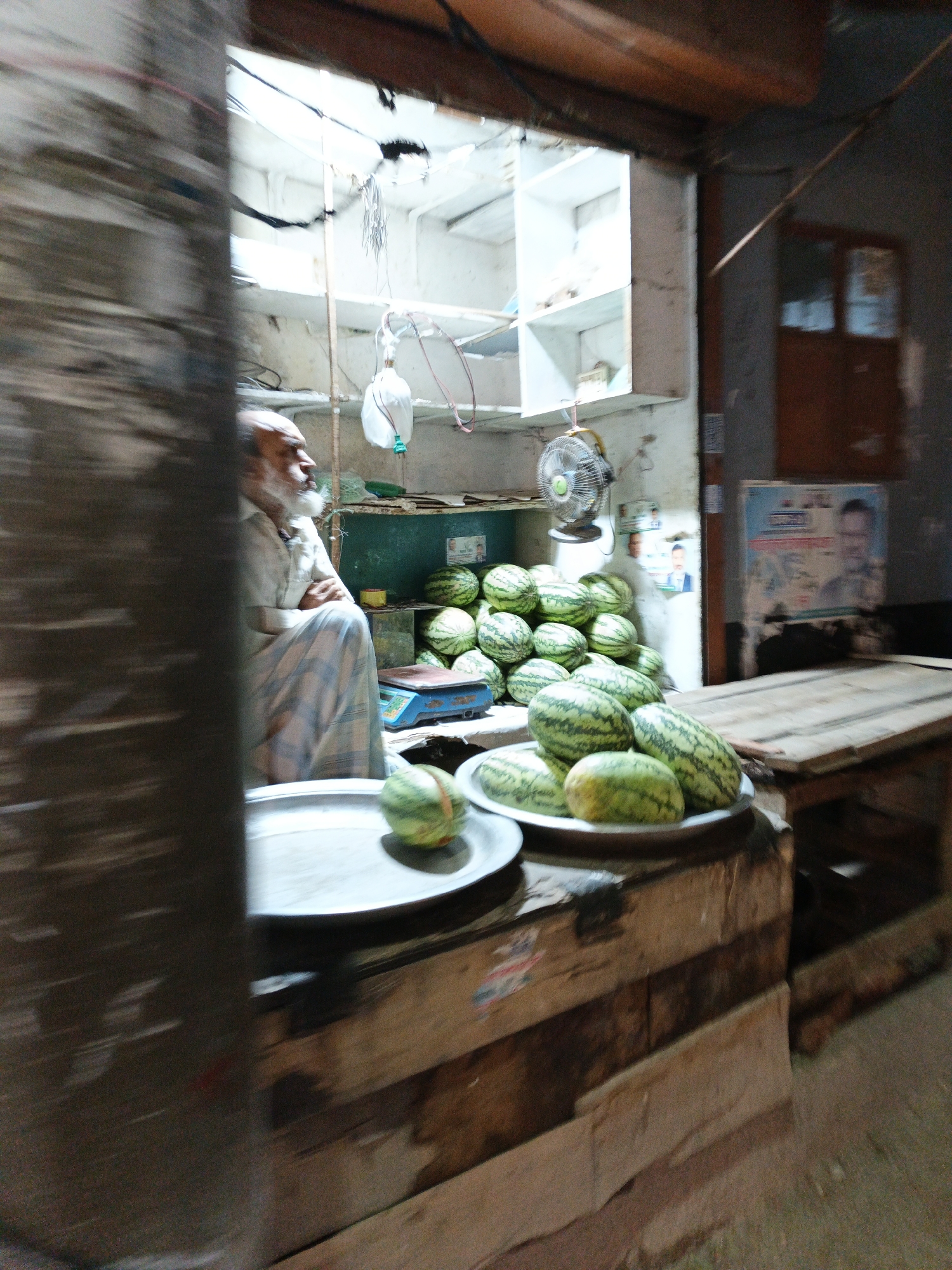 An eye-level, blurred photograph depicts a watermelon stand in a shadowed area. A bearded man wearing a plaid garment sits behind a stack of green watermelons in the background of the frame. A small fan is seen on the back wall above the watermelons. There are a number of cables, wires, and tubing attached to the upper section of the interior space. In the foreground are two metal trays filled with the same watermelons. The surfaces the trays sit on are made of a dark stained wood with paint. A wooden table is seen on the right side.