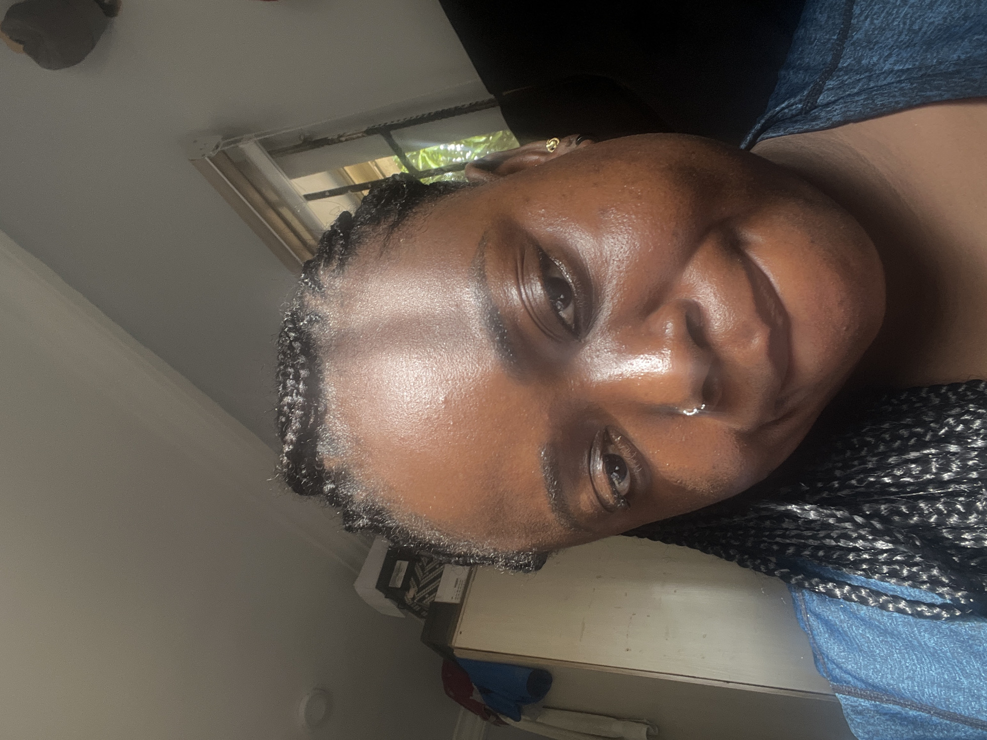 A close-up, tilted portrait captures a person with dark skin and dark, box-braided hair, looking directly at the viewer with a slight smile. The individual's face is illuminated, highlighting a natural sheen on their forehead and cheeks. They are wearing a small silver nose ring on their right nostril and a tiny gold hoop earring in their right ear. A dark blue or grey textured top is visible on their upper body.
The background suggests an indoor setting. A window is partially visible behind the person's head, showing green foliage outside, indicating natural light and an outdoor environment with vegetation. The upper part of the image features a plain, light-colored wall or ceiling. Towards the bottom right, a light-colored wooden cabinet or table is seen, with a blue fabric item and other indistinct objects on it. The image appears to be a selfie or a casual close-up taken during the day. No text is visible. The location context provided is Kubwa, Nigeria.