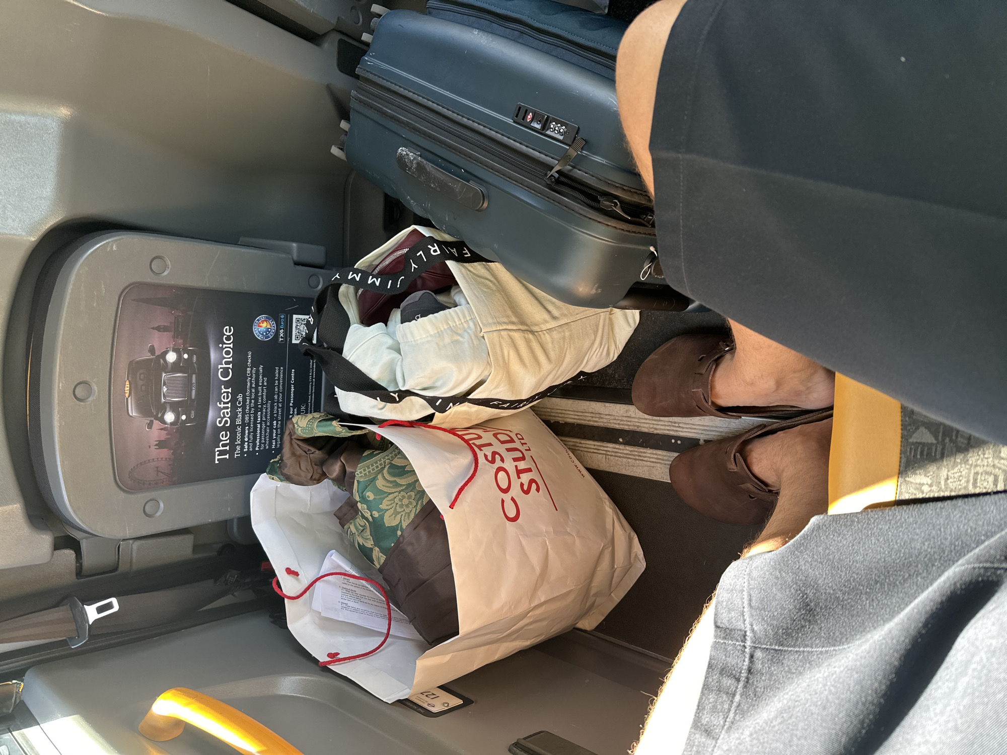 The image shows luggage and shopping bags piled on the floor of a London black cab, next to the seat where a person is sitting. One luggage item is a blue-grey suitcase, and two open shopping bags labeled "COSTU STUDIO LTD" contain assorted cloth items and clothing. The passenger's legs and brown shoes are visible. A poster about the "Safer Choice" of London black cabs is visible on the partition of the cab.