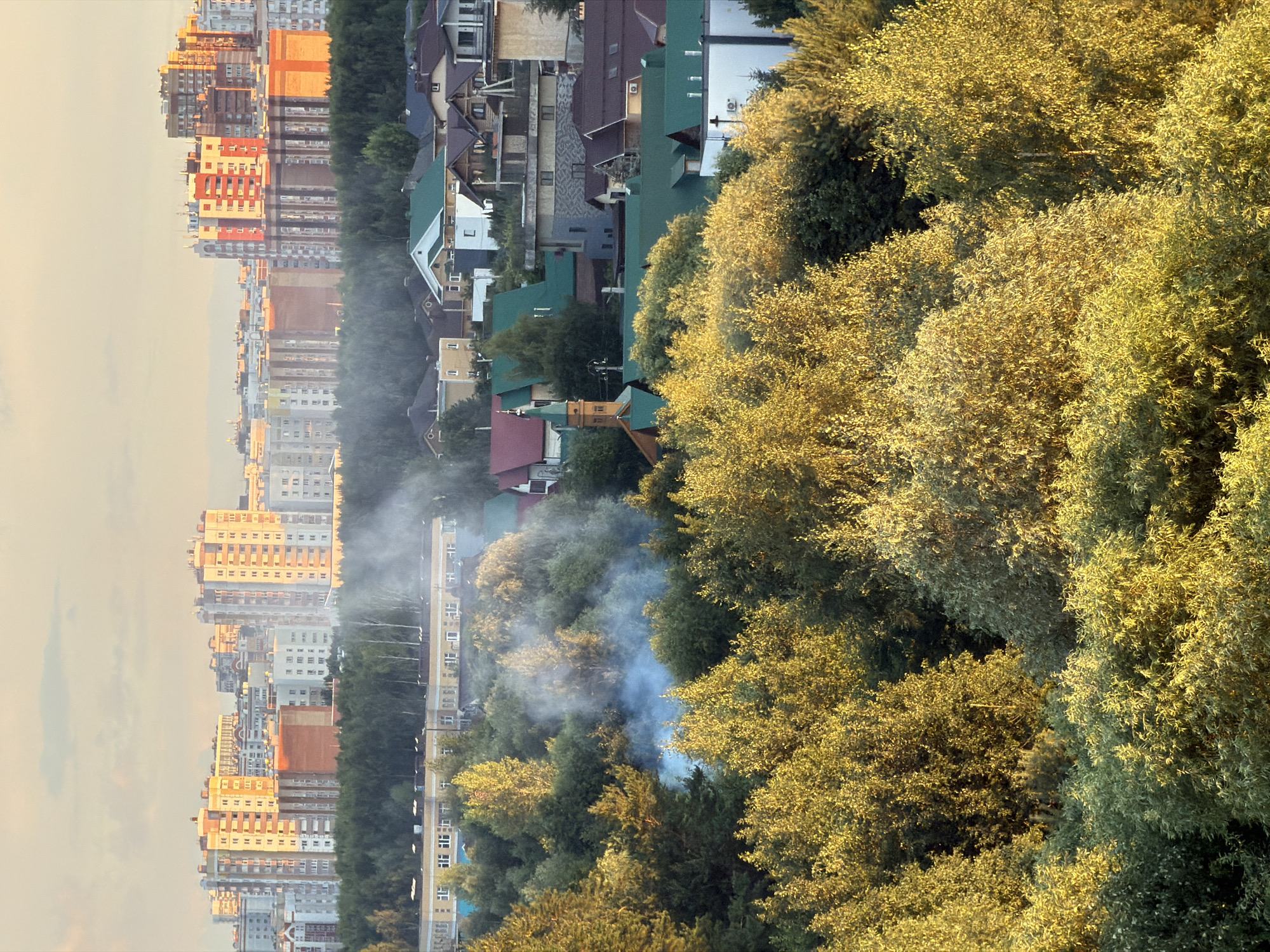 An elevated medium shot captures a suburban cityscape under a soft, hazy sky, likely during sunrise or sunset, given the warm golden light illuminating the scene. In the foreground, a dense canopy of lush green trees, their leaves catching the light and appearing golden and yellow, dominates the lower half of the frame. Some trees have a lighter, more yellow hue, suggesting birch or aspen. In the mid-ground, a plume of light blue-gray smoke rises from within the trees, obscuring parts of the foliage and suggesting a fire or controlled burn. Behind the trees, a strip of darker green forest forms a transition to the background.
Further in the background, a collection of low-rise residential buildings with diverse roof colors, predominantly green, brown, and gray, is visible. Some have pitched roofs, while others are flatter. A few buildings feature white walls and darker trim. A distinct building with a green steeple and a pointed roof, resembling a church or chapel, stands out among the houses. Beyond the residential area, a line of taller, multi-story apartment buildings forms the skyline. These buildings are constructed from materials in shades of beige, white, and orange, with many windows visible. The sky above is a pale, yellowish-pink, with subtle cloud formations. The overall impression is of a developed area with a significant presence of natural greenery.