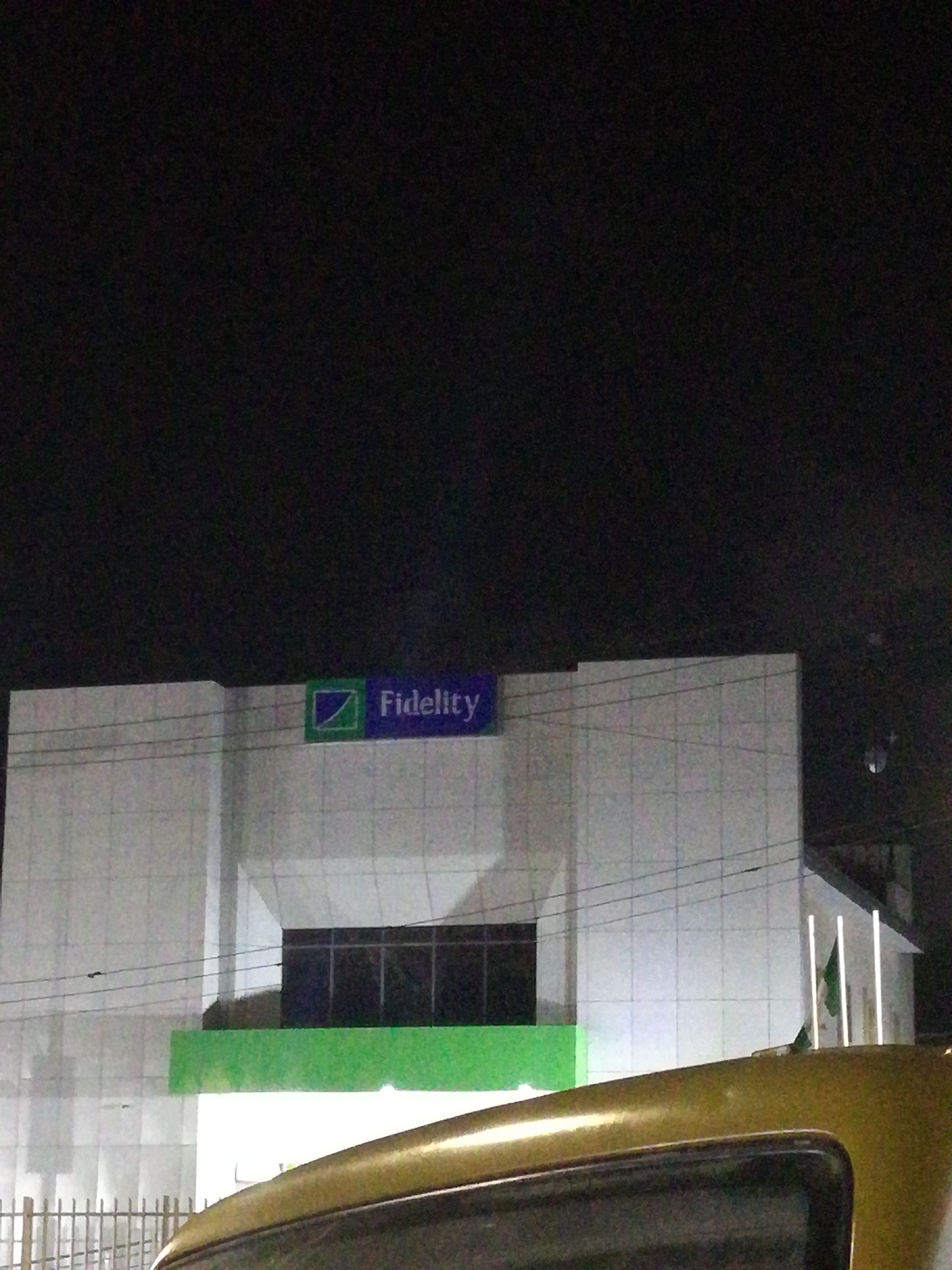 The image shows a Fidelity Bank building at night. The building is white with a green stripe running along the lower level and a blue sign on top that reads "Fidelity" with the bank's logo. The sky is dark. A vehicle appears in the foreground. The image was taken in Lagos, Nigeria. There are no visible people.