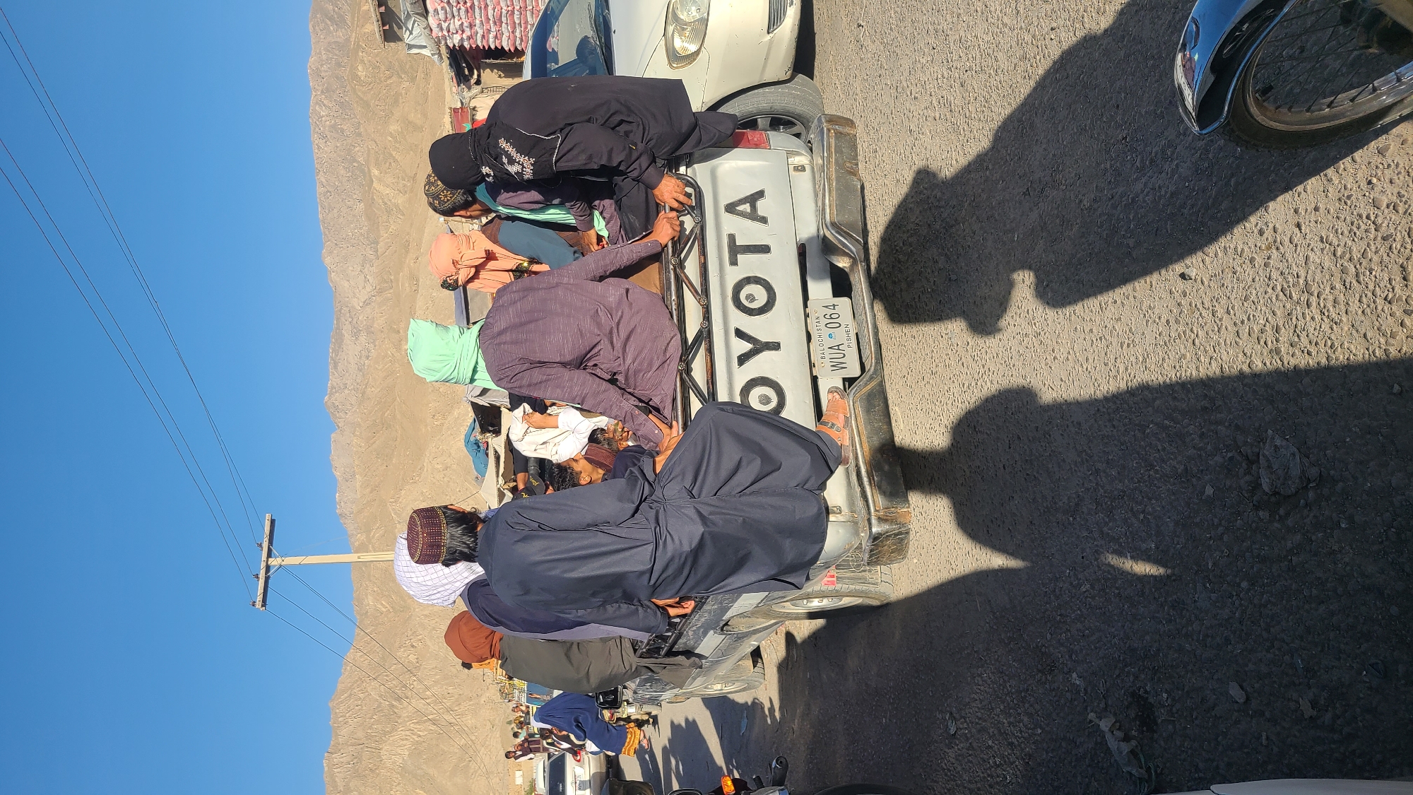 The image depicts a group of individuals riding in the back of a silver Toyota pickup truck. The truck's license plate reads "WUA 064" and indicates it is from the Baluchistan region of Pakistan. The people in the truck are predominantly dressed in traditional attire, including dark-colored robes and head coverings. Some are positioned near the front of the truck bed, leaning towards the cab. The setting appears to be a roadside in a mountainous area, with a clear blue sky above. A utility pole with wires is visible in the background, and other vehicles and people are nearby. The location is identified as Kuchlak, Pakistan. The sun casts shadows on the road, indicating the time of day.