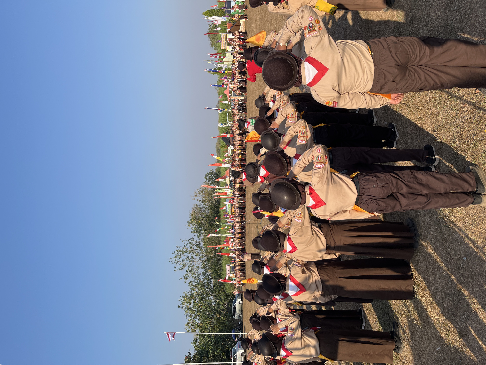 The image depicts a scene on a dry, grassy field under a clear blue sky, likely in the city of Gembong, Indonesia, given the context. A group of individuals, predominantly young people dressed in scout uniforms, are gathered. The uniforms consist of tan shirts, brown skirts or trousers, red neckerchiefs, and dark hats. Many of them are shown from the back or side, with their right hands raised in a salute. They appear to be facing a group further in the distance, where more scouts and several flags are visible. The flags are of various colors and designs, suggesting a formal event or gathering. In the background, trees and some structures are present, along with parked vehicles. The sunlight casts long shadows across the field, indicating it is likely morning or late afternoon.