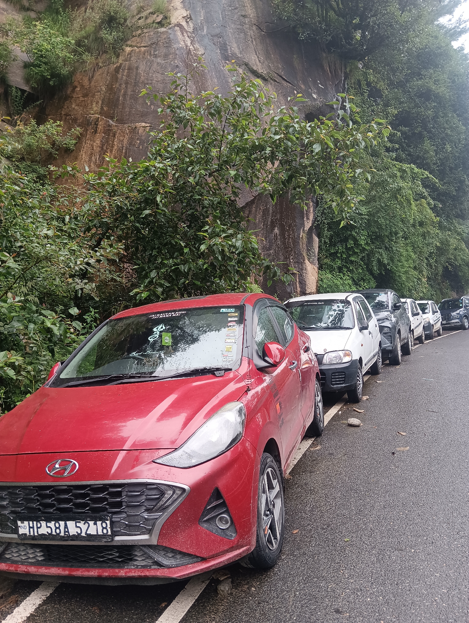 A red Hyundai car, license plate HP 58A 5218, is parked on the right side of a wet, asphalt road next to a steep, rocky cliff face overgrown with green foliage. A line of other cars, predominantly white and black, are also parked along the roadside, extending into the distance. The environment appears to be a natural landscape, possibly a mountainous or hilly region. The sky is overcast, contributing to the diffused lighting. No people are visible in the frame. The car has various stickers on its windshield and exterior.