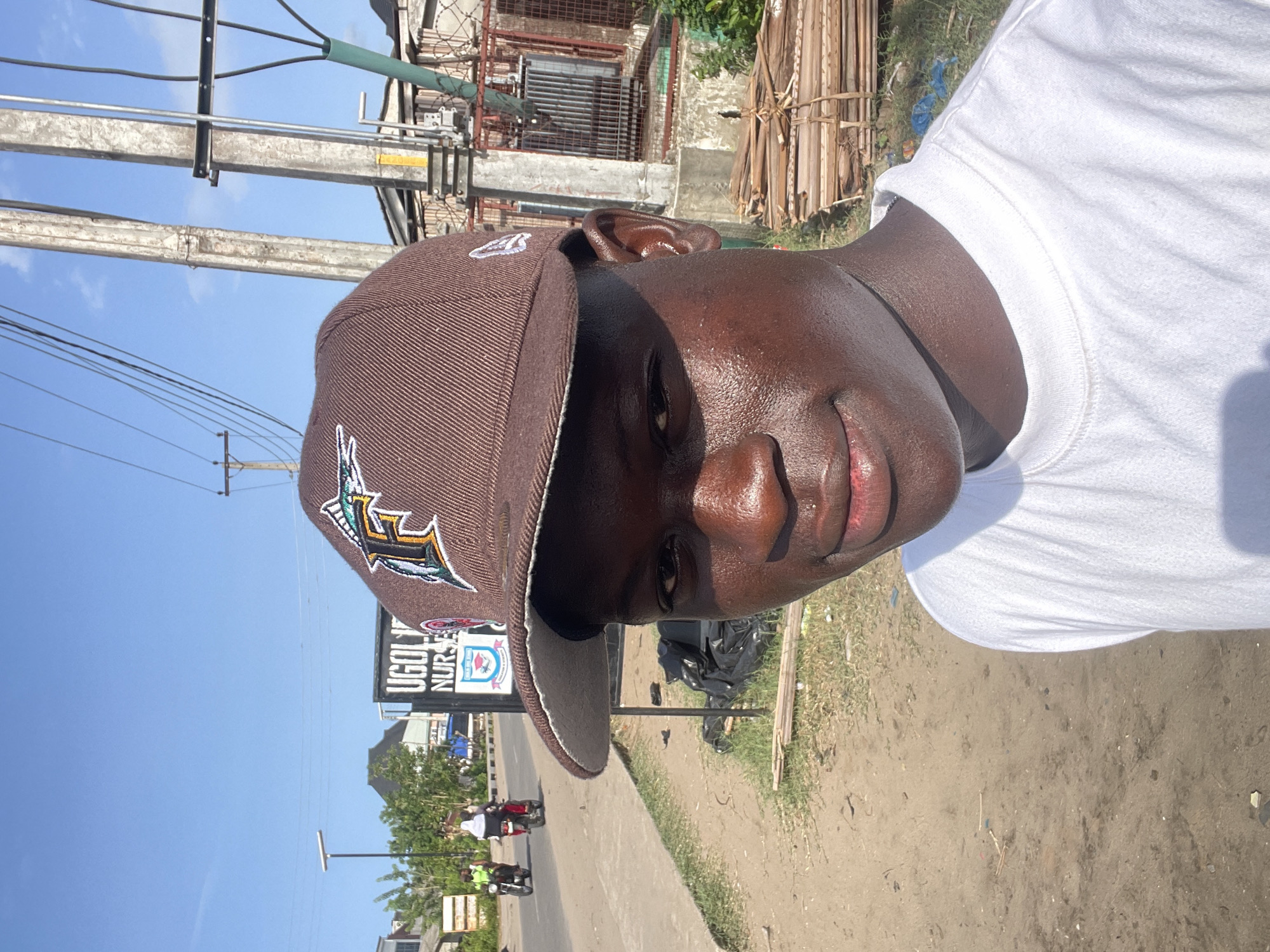A close-up, rotated image captures a person wearing a brown baseball cap with a sports logo and a white t-shirt. The individual's skin appears oily or sweaty.
The background reveals an urban roadside setting in what appears to be Badagry, Nigeria. A sign reading "UGOLI NUR..." suggests the presence of a medical facility or pharmacy in the area. Power lines are visible overhead. A dirt shoulder borders the asphalt road, where two motorcycles are moving, and litter is scattered. Construction materials lie adjacent to an adjacent building.