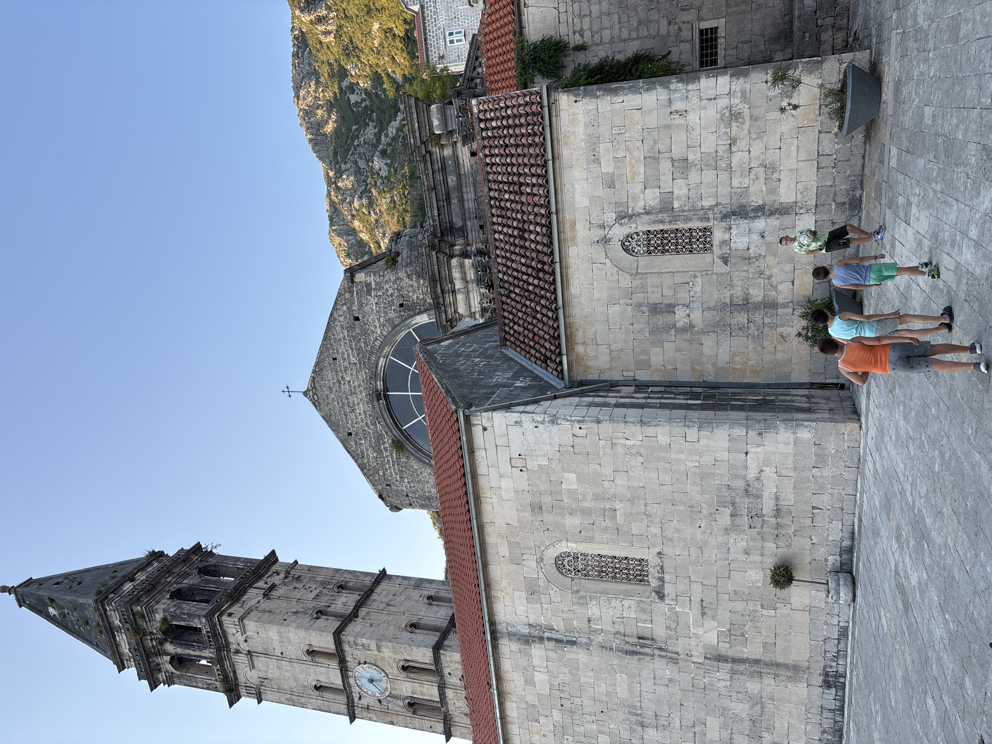 In the city of Perast, Montenegro, a stone church and its bell tower dominate the frame under a clear blue sky. The bell tower, to the left, features a clock with Roman numerals and a pointed, weathered spire. Adjacent to it, the main church structure displays a large, arched window and a red-tiled roof. The facade is constructed of large, light-colored stone blocks. In the foreground, four young boys are walking away from the camera, dressed in casual summer clothing. One boy, slightly further back and to the right, stands holding a ball, observing the group. A single, potted tree with a rounded top is visible near the left side of the church wall. The ground is paved with grey, square stones. In the background, a lush, green mountainside rises, bathed in sunlight.