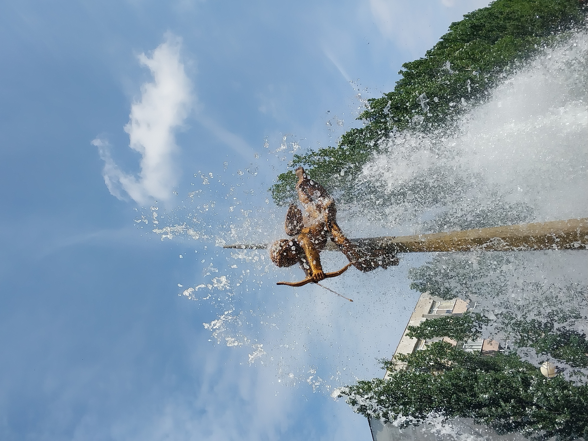 The image captures a golden statue of Cupid, complete with bow and arrow, adorned with a deluge of water from an active fountain. The statue is centrally positioned against a backdrop of a clear blue sky dotted with white clouds. To the left, a multi-story building stands behind a row of lush green trees. To the right, more green trees form a dense line. The fountain's spray is dynamic, with numerous water droplets in motion, obscuring parts of the statue and the surrounding foliage. This fountain is located in the city of Dnipro, Ukraine, specifically in the area of Pobeda Park.