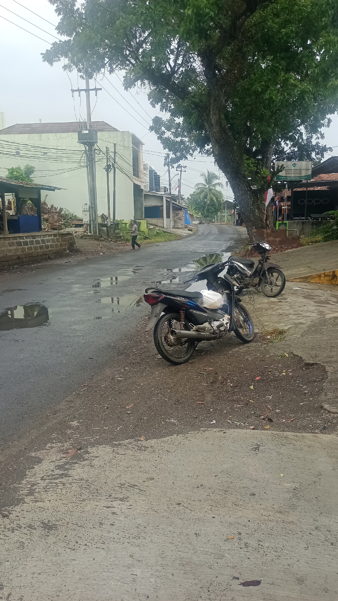 The image depicts a street scene in Cikalong, Indonesia, on an overcast day. The road is wet and reflects puddles of water, suggesting recent rain. In the foreground, two motorcycles are parked on the side of the road. The closest motorcycle is black with blue accents and has a white bag on its seat. The second motorcycle, parked slightly behind and to the right, is a darker color and appears older.
The environment includes a tree with dense green foliage on the right side of the frame, partially obscuring a building. The building on the right has a sign with green and white branding, possibly a business, and an "OPPO" logo is visible. Flags, including an Indonesian flag, are displayed near this building. On the left side of the street, there are buildings with light-colored walls, some appearing to be residential or commercial structures. A power pole with transformers and overhead wires is prominent on the left. A lone individual is walking away from the camera on the road in the middle ground. The overall impression is a somewhat weathered urban or semi-urban environment.