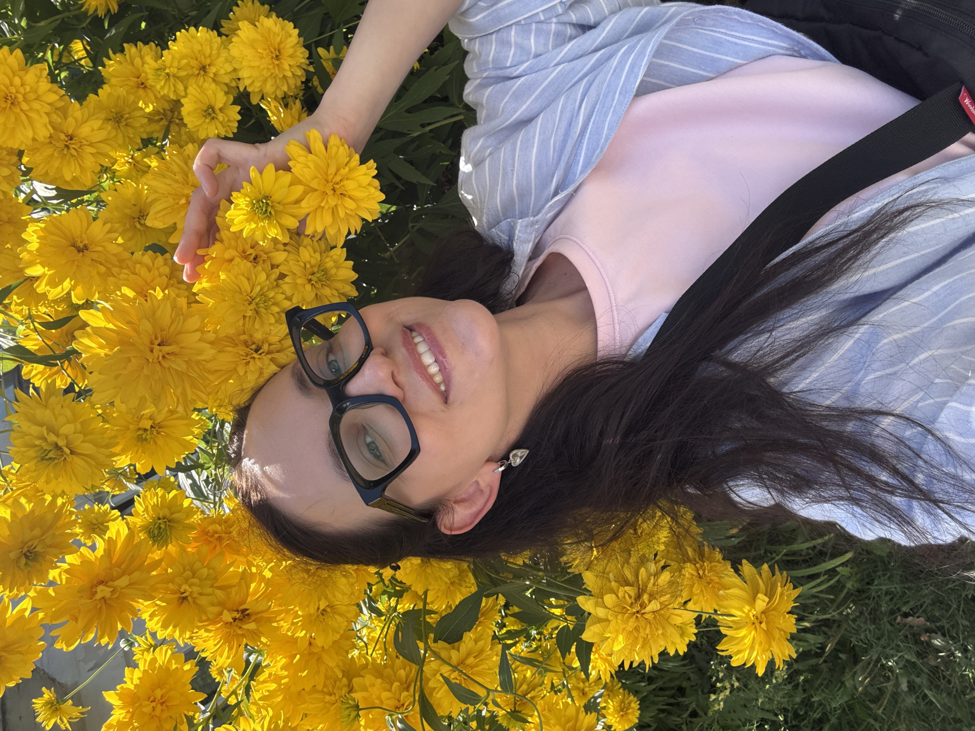 A young woman with dark hair and black-rimmed glasses is lying down amongst a cluster of bright yellow flowers. She is wearing a light pink t-shirt and a light blue and white striped button-up shirt, with a black strap crossing her chest. Her right hand is raised, holding one of the yellow flowers near her face. The flowers are in full bloom and create a vibrant yellow backdrop for the woman. The surrounding environment appears to be an outdoor setting, possibly a garden or park. The image is taken from a high-angle perspective. It is not possible to determine the specific location in Chaikovsky, Russia, from the visual information provided.