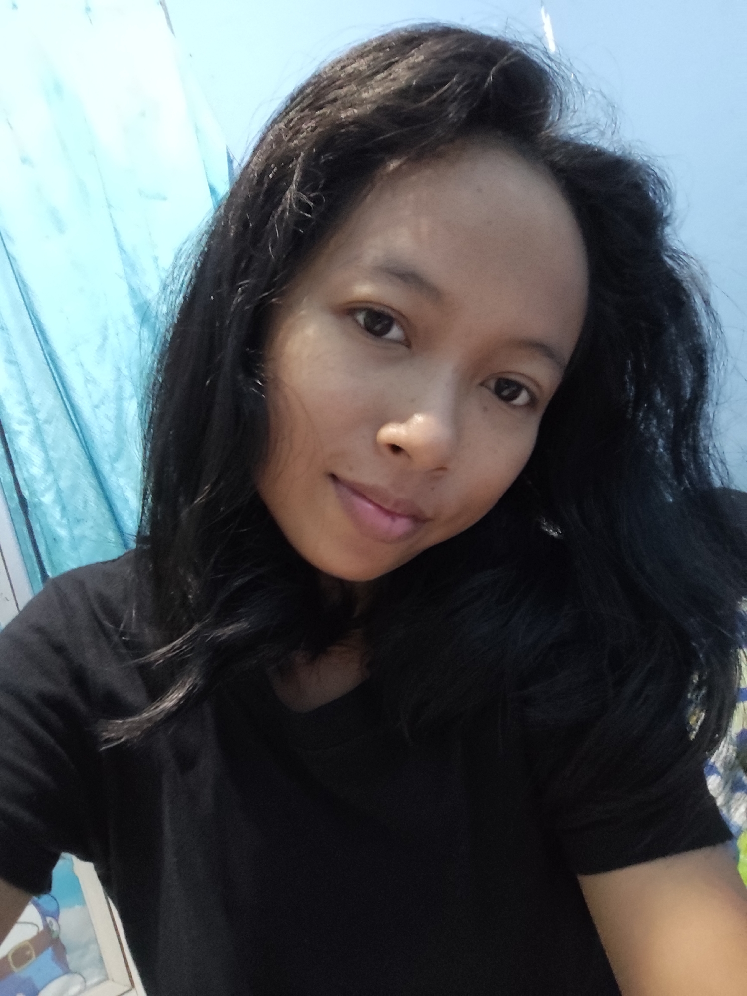 A young woman with dark, wavy hair is the subject of this close-up, selfie-style photo. She is wearing a plain black t-shirt. Her skin tone appears to be light brown. Her expression is neutral, and she is looking slightly off-camera. The background consists of a light blue curtain or fabric on the left side of the image, and a lighter blue wall or surface on the right. There are some indistinct patterns or objects visible in the lower right corner, possibly part of a blanket or bedding. The lighting suggests it is daytime, but no specific time of day is discernible. There is no visible text in the image.