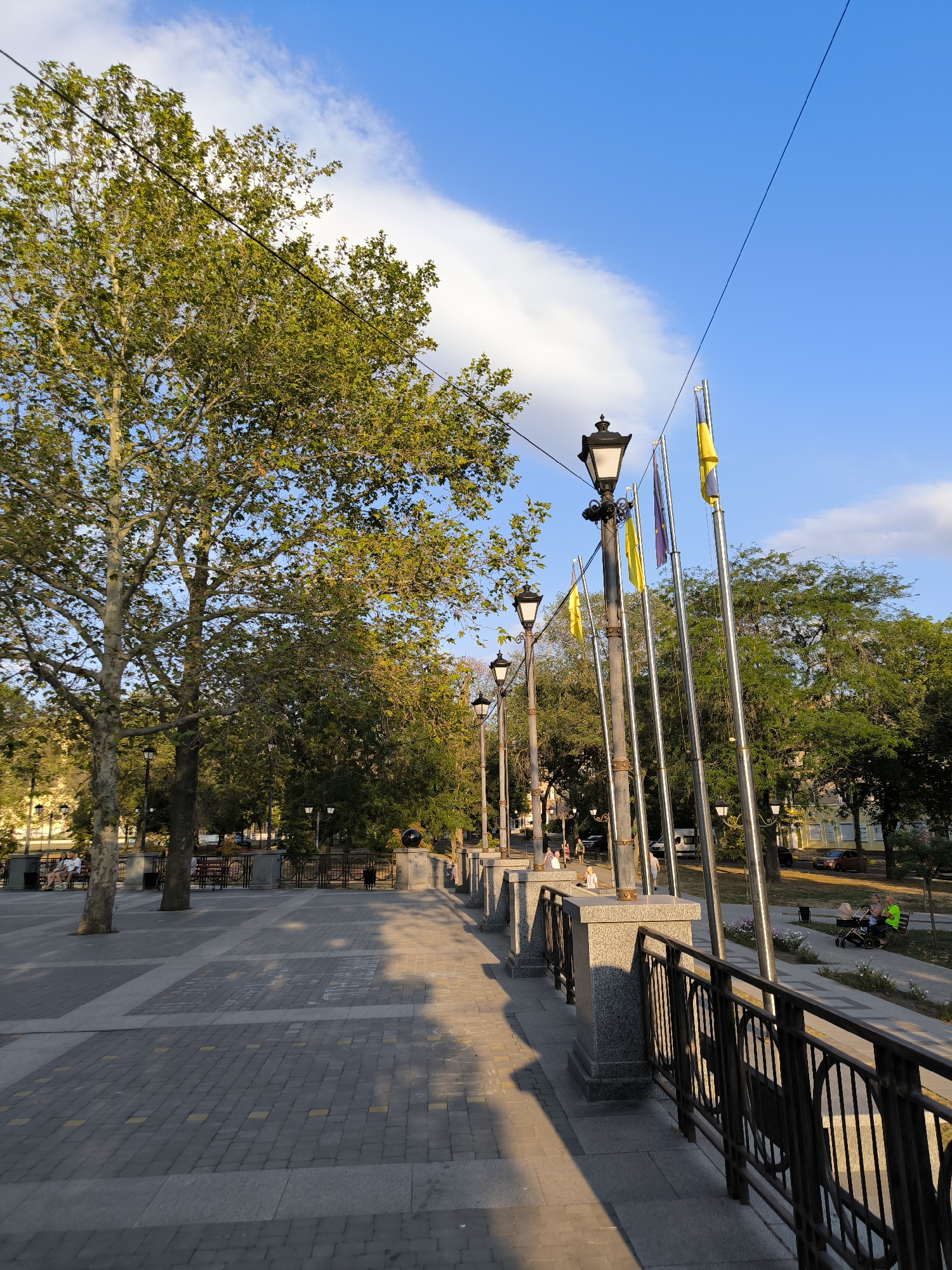 The image depicts a paved walkway in a park-like setting, likely in Mykolaiv, Ukraine, given the presence of Ukrainian flags. The walkway is lined with lampposts and a metal railing. Large trees with green foliage are visible on both sides of the walkway. In the background, there are more trees, buildings, and a few people are seated on benches. The sky is blue with some white clouds, and the sun is casting long shadows, suggesting it is either early morning or late afternoon. The overall scene conveys a peaceful, public outdoor space.