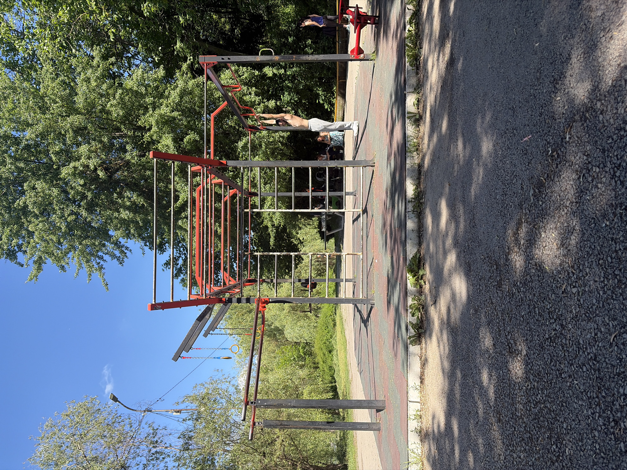 The image shows an outdoor exercise area in Targovishte, Bulgaria. A man is performing a horizontal exercise on monkey bars. Another person is on exercise equipment in the background. The area has trees and a walkway.