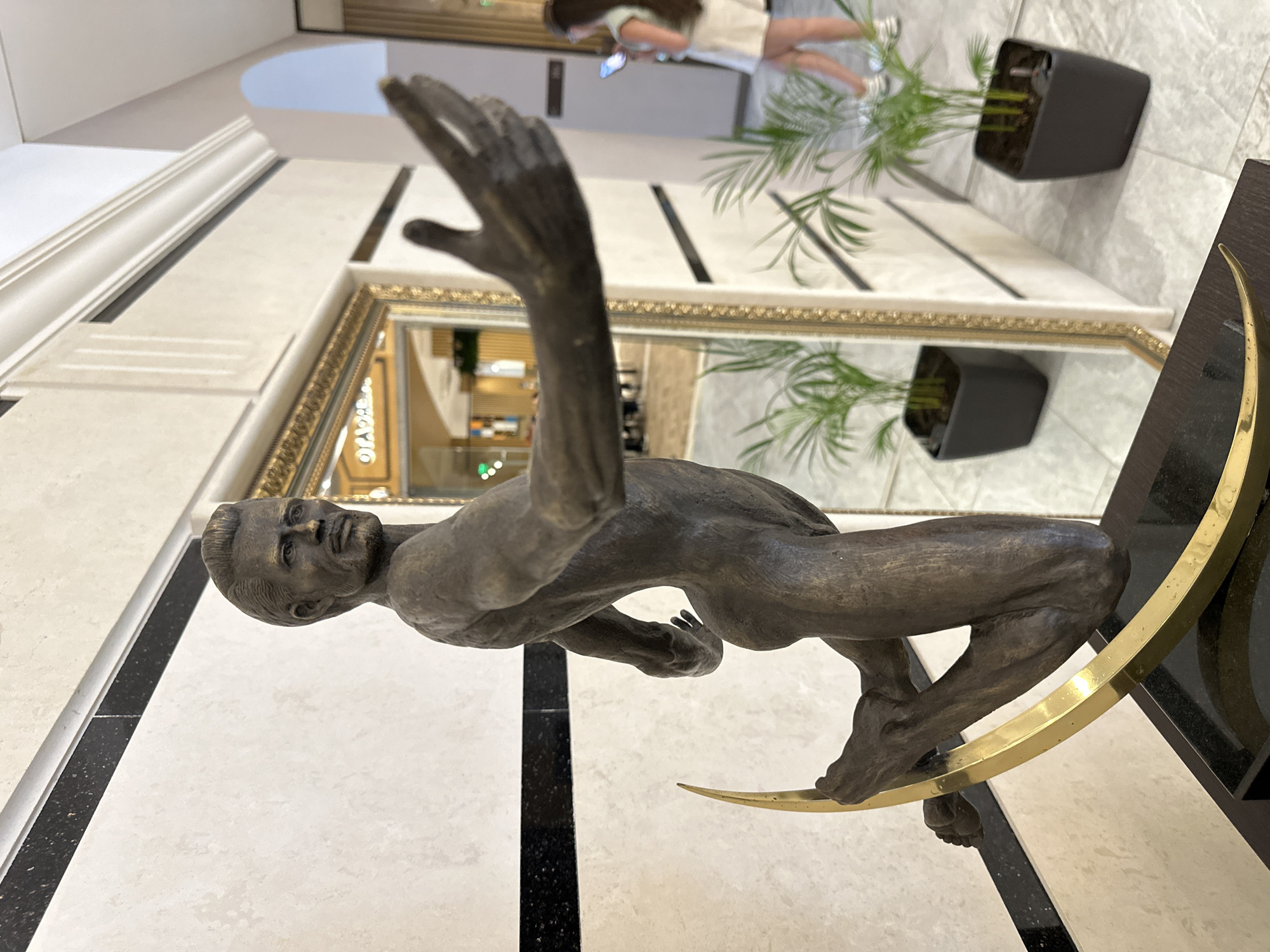 An eye-level indoor shot in Razliv, Bulgaria, features a bronze sculpture of a male figure in a dynamic, balanced pose, facing the left of the frame. The muscular, nude figure has one arm extended upwards and the other bent, seemingly poised on a polished golden crescent-shaped stand. The sculpture's face, visible in profile, shows a serious expression.
The sculpture is set on a dark, flat surface. The background reveals a brightly lit interior space with polished floor tiles, predominantly light-colored with striking black stripes. A large mirror with an ornate gold frame occupies a significant portion of the background, reflecting parts of the room, including a potted plant with green leaves in a dark planter. In the upper left background, part of a wall with white molding is visible.
Through the reflection in the mirror, and partially in the actual background, a woman is seen. She wears a light-colored top and skirt and appears to be looking at her phone. The general ambiance is modern and clean, characteristic of a hotel lobby or a public building. The lighting is bright and even, typical of an indoor setting, and no specific time of day is discernible. There is no visible text.