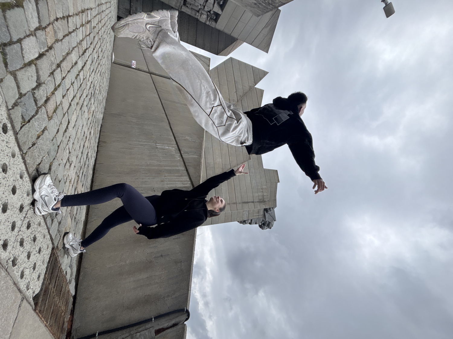 This image, intentionally rotated, captures two individuals in dynamic, seemingly gravity-defying poses against a backdrop of stark architecture and a cloudy sky. A young woman, positioned on what appears to be a vertical cobblestone-paved surface, wears a black hoodie, dark blue leggings, and white athletic shoes. She extends her right arm upwards and outwards, looking towards the other person. Above and to her right, another individual, likely male, is captured in mid-air, horizontally suspended. This person wears a black hoodie and light-colored, loose-fitting pants adorned with geometric black outlines, along with white athletic shoes. Their left arm is also outstretched, creating an impression of reaching towards the first individual, as if in a dance or a cinematic moment of connection or separation.
The setting features large, angular concrete structures that suggest a modern architectural complex or a brutalist monument, contributing to the dramatic and somewhat surreal atmosphere. The perspective is from a low angle, looking up towards the sky, which is overcast with thick gray clouds. The lighting is diffused, indicating daytime with no direct sunlight. The ground or wall to the left is textured with large, irregular paver blocks or cobblestones, adding visual interest. The overall impression is a staged, artistic photograph playing with perspective and illusion. No discernible text is visible in the image.