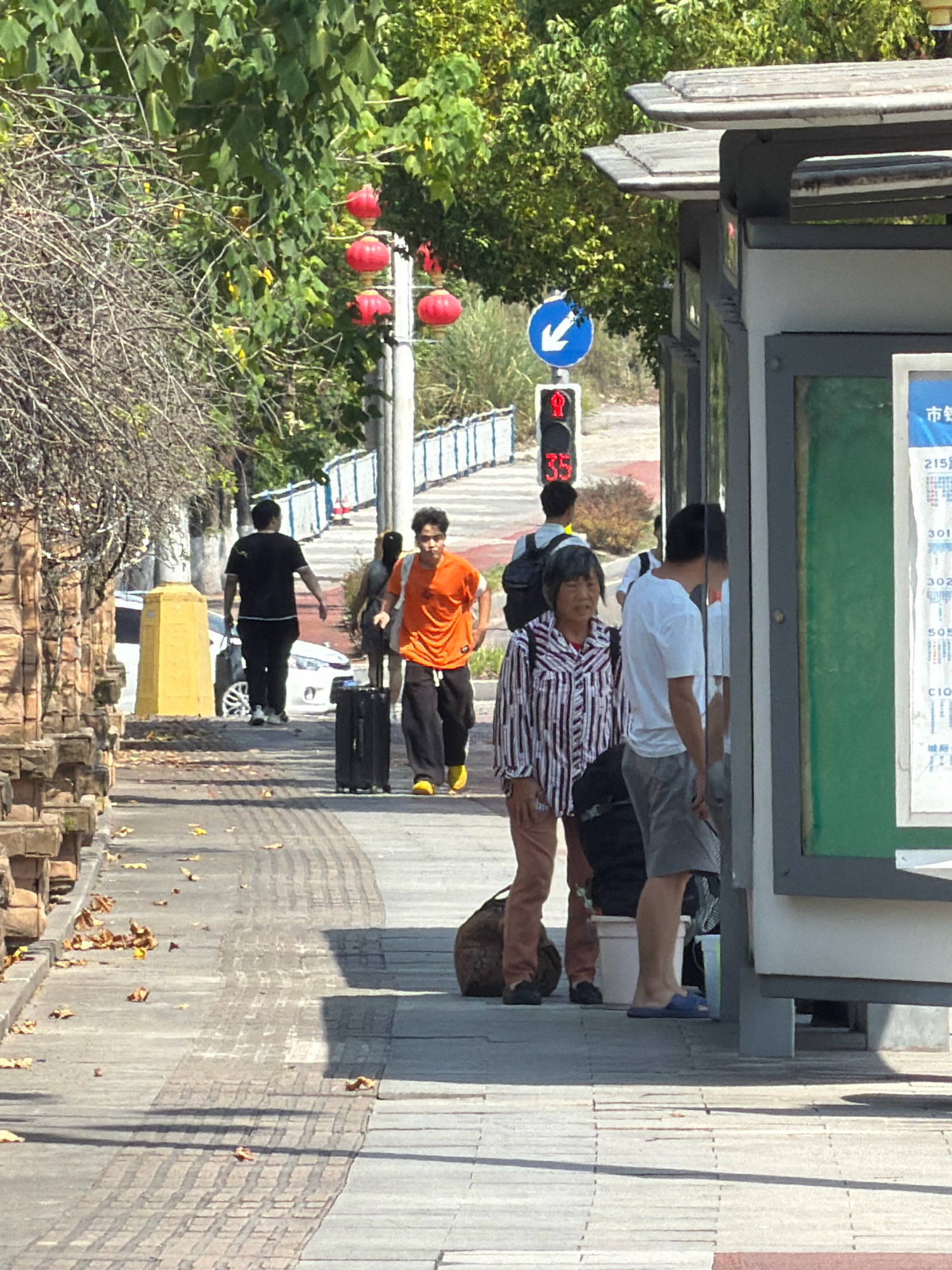 A sunny day scene captures a street in the city of Jiangjin, China, with a bus stop in the foreground. A group of people are waiting or walking along a sidewalk adjacent to a road. In the immediate foreground, a bus shelter stands to the right, displaying a route map. To the left of the bus shelter, a woman in a striped shirt and a man in a white t-shirt and shorts are visible. Further down the sidewalk, a young man in an orange t-shirt is walking with a black suitcase, and another person in a black t-shirt is walking away from the camera. A traffic light displays a red signal and the number "35". Red lanterns hang from a pole in the background, suggesting a festive or decorative element. The environment includes trees and foliage, and the ground is paved with concrete and patterned tiles.