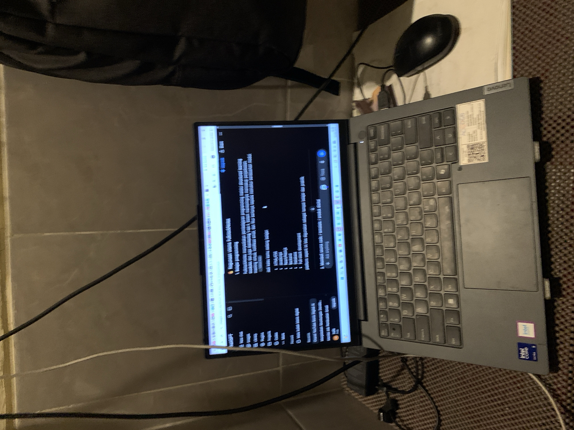 In an indoor setting, a laptop is open and displaying a webpage with text in Indonesian, likely on a desk or table. The laptop's screen shows a website titled "FullstackNotes" with sections such as "Belajar programming" (Learn programming) and "Apa yang ingin kamu pelajari?" (What do you want to learn?). The user interface suggests a learning platform or educational resource. A black computer mouse is positioned to the right of the laptop. The environment appears to be dimly lit, with power cords and cables visible. The wall in the background is textured and light brown. The image also shows a portion of a black backpack or bag on the right side of the frame. In the bottom left of the laptop, there are "Intel Core Ultra" stickers. The overall impression is a workspace with technology, likely used for learning or development.