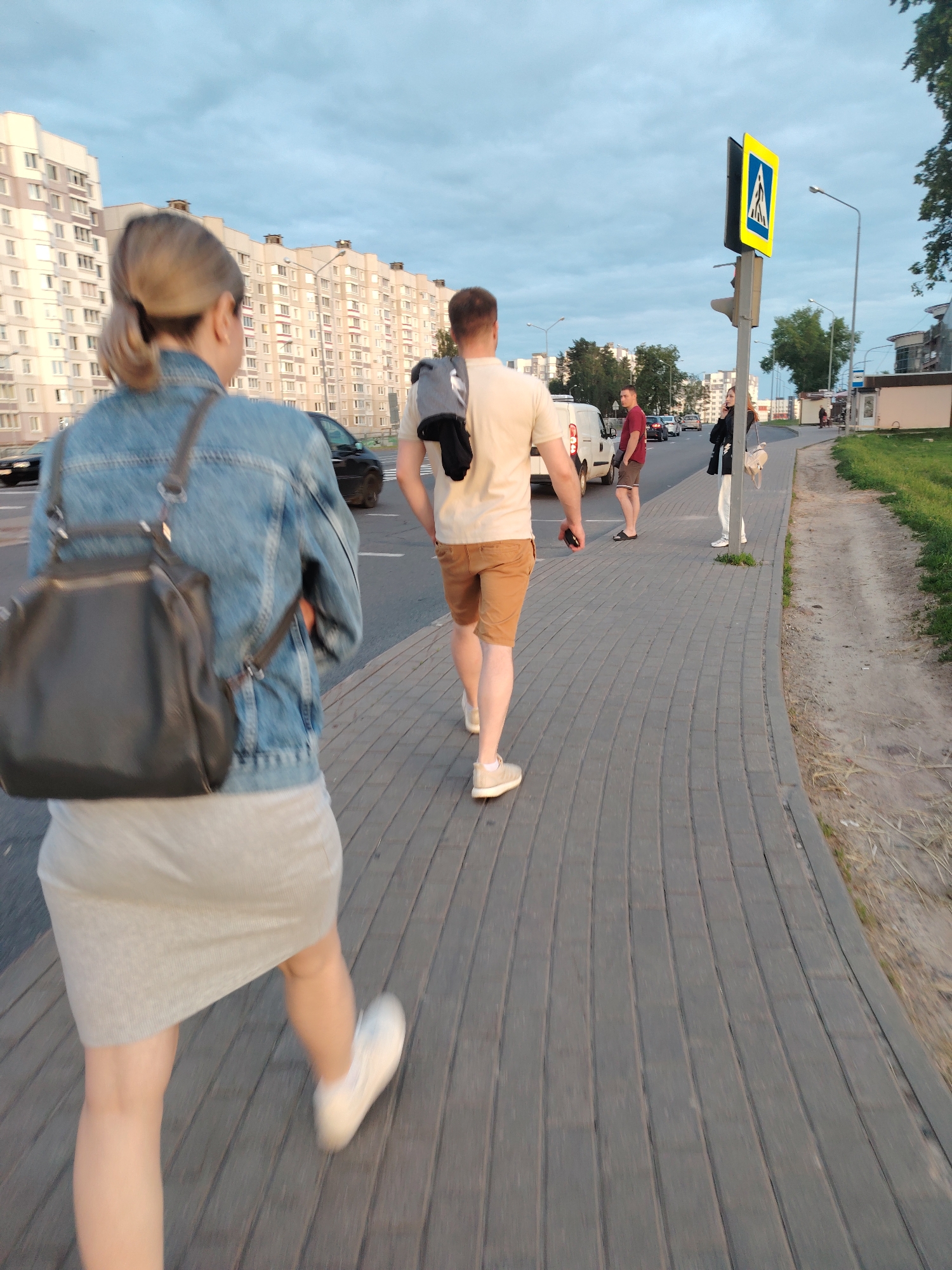 The image depicts an outdoor urban scene in the city of Бараўлянскі сельскі Савет, Belarus, under an overcast sky, likely in late afternoon or early evening.
In the immediate foreground, a female subject walks away from the viewer on a grey paved sidewalk. She has light-colored hair styled in a low ponytail, wears a blue denim jacket over a light grey ribbed midi skirt, and white sneakers. A dark grey or black leather backpack is worn over her shoulders.
To her right, a male subject also walks away from the viewer on the same sidewalk. He has short, light brown hair, wears a light beige t-shirt, brown shorts, and light beige sneakers. A grey item of clothing or a bag is draped over his left shoulder, and he holds a dark object in his right hand.
Further down the sidewalk, on the left side of the image, a second male subject stands next to a white panel van parked on the edge of the asphalt road. This male wears a red t-shirt, dark shorts, and dark flip-flops, and faces towards the left of the frame.
Adjacent to him, a second female subject stands on the sidewalk, facing towards the left, with a phone held to her right ear. She has dark hair, wears a black jacket, white pants, and carries a light beige handbag.
A yellow pedestrian crossing sign, featuring a white pedestrian icon within a blue square, is visible on a black pole behind the second female subject. A dark traffic light is mounted below the sign, and a tall, thin grey street light pole extends upwards.
The sidewalk is constructed from grey interlocking pavers arranged in a curved pattern. To the right of the sidewalk, a narrow, unpaved dirt path with sparse grass runs parallel to it.
The asphalt road, partially visible, displays white lane markings. Several vehicles are present on the road, including a black car behind the foreground female and other cars in the distance.
In the background, a series of multi-story residential apartment buildings with light-colored facades (beige and white) dominate the skyline. Scattered green trees are visible between and beyond the buildings. The sky is largely overcast with light blue-grey clouds.