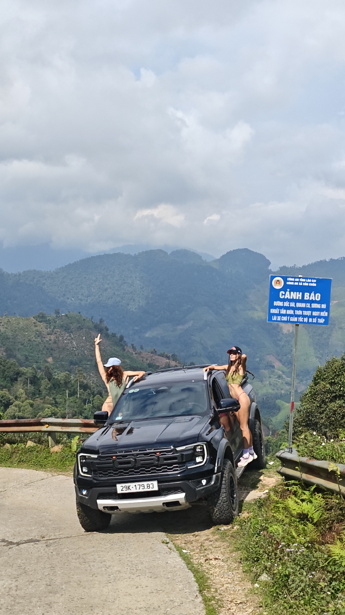 Two women in swimwear stand on and around a black Ford truck parked on a narrow road in a mountainous region. The truck has a license plate that reads "29K-179.83". One woman is standing on the truck's roof, gesturing with her arm raised. The other woman is standing on the truck's step, near the open door. Behind them, a blue road sign with white text in Vietnamese is visible. The sign reads "CẢNH BÁO", which translates to "Warning". The background shows a lush, green, mountainous landscape under a cloudy sky. This image was taken near Suối Giàng, Vietnam.