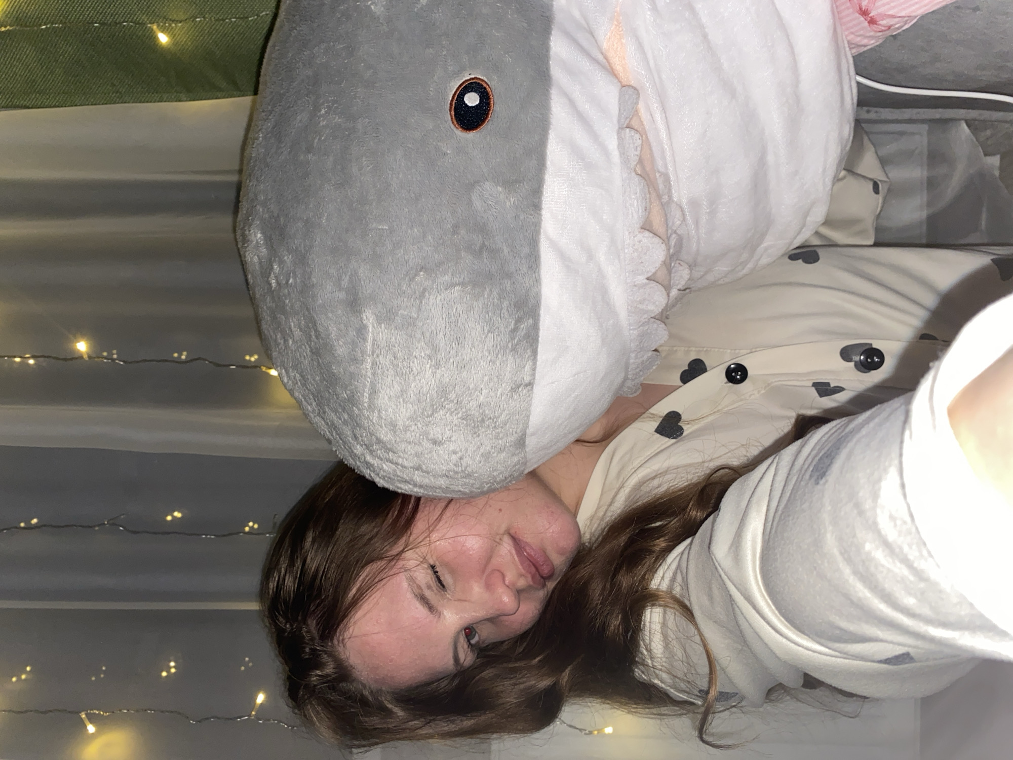 A woman lies on her back, indoors, with a large plush shark toy resting on her forehead. She has brown hair and is wearing pajamas with a heart pattern. The background includes soft lighting from fairy lights and possibly a green cushion or blanket. There is no verifiable information to suggest this image was taken in Izhevsk, Russia.