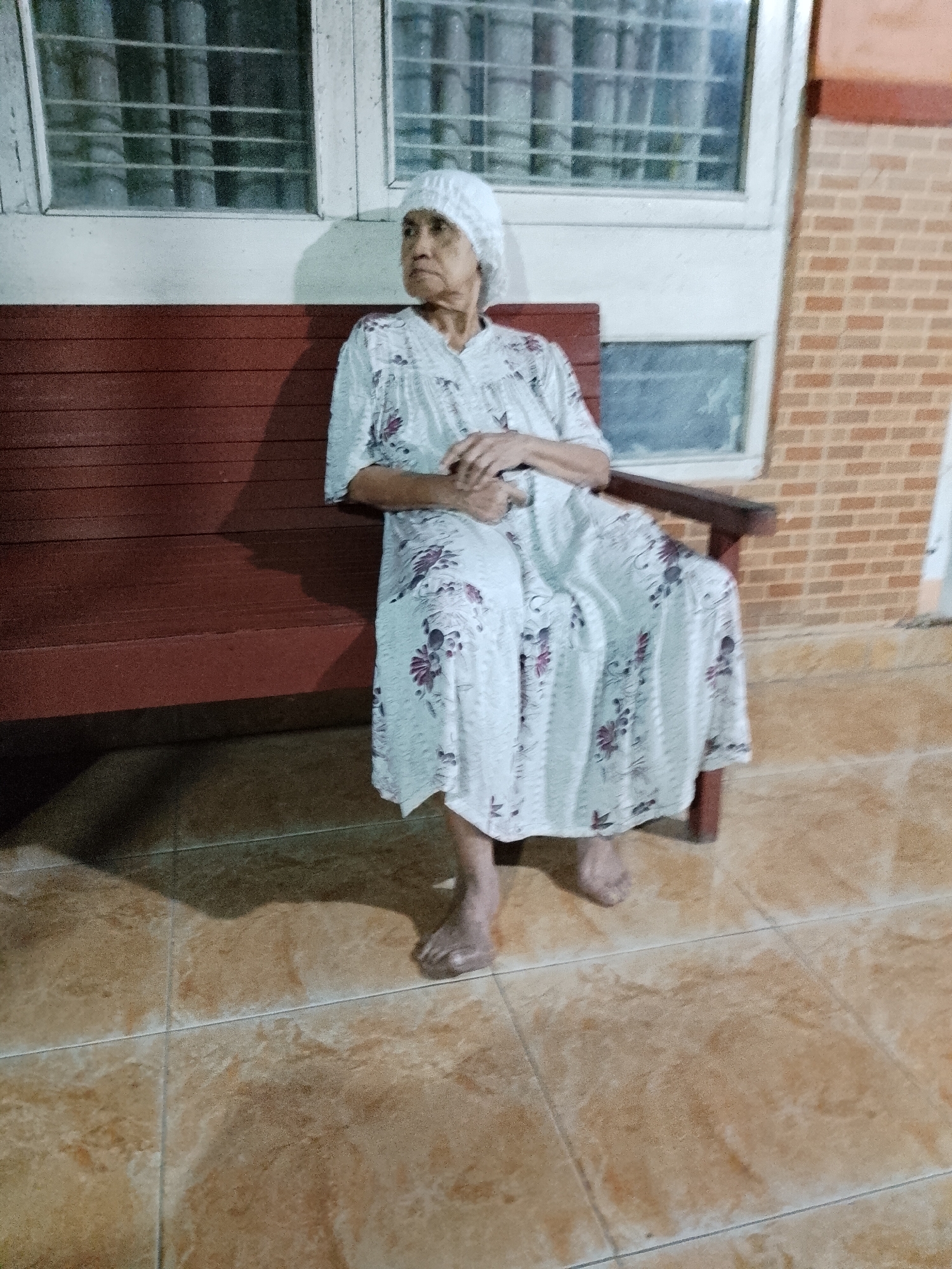 An elderly woman, wearing a white head covering and a light-colored, floral-patterned dress, sits on a dark red wooden bench. She is looking to her left, with her hands clasped in her lap. The bench is positioned on a tiled outdoor patio with tan, marble-patterned tiles. Behind the bench, there is a white-painted wall with two windows that have visible metal grates. To the right of the wall, there is a section of brickwork. The lighting suggests it is either early morning or late evening, as indicated by the shadows cast on the floor. The scene appears to be in a residential setting in Kecamatan Pati, Indonesia.