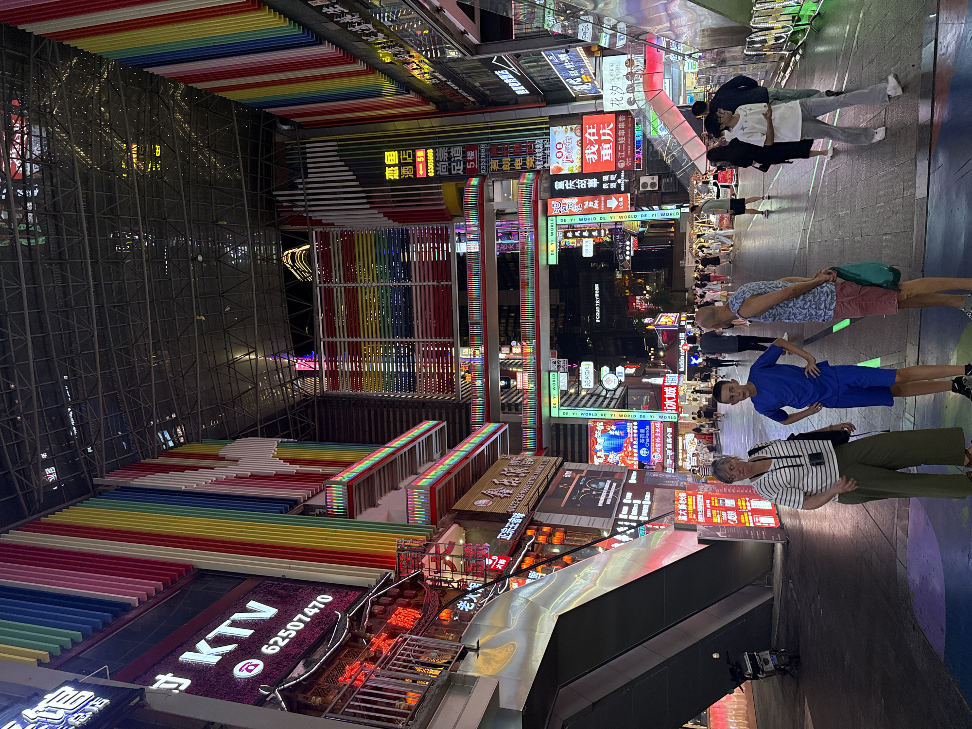The image depicts a nighttime scene in Yuzhong District, China, specifically in a vibrant, pedestrianized area illuminated by abundant neon signage and colorful architectural lighting. In the foreground, a woman wearing a striped t-shirt and green pants stands to the left, facing forward with her head slightly turned. To her right, a young boy in a blue t-shirt and shorts also faces forward. Behind them, other pedestrians are visible walking in the illuminated thoroughfare, which is lined with multi-story buildings adorned with numerous signs in Chinese characters, some featuring images and phone numbers. The architecture is characterized by a mix of modern and traditional elements, with exposed metal structures overhead and colorful vertical ribbed facades on the buildings. Escalators and staircases are integrated into the buildings, also lit with colored lights. A scooter is parked to the left, near a large, angled escalator. The overall atmosphere is bustling and commercial, with a strong emphasis on visual stimulation from the lights.