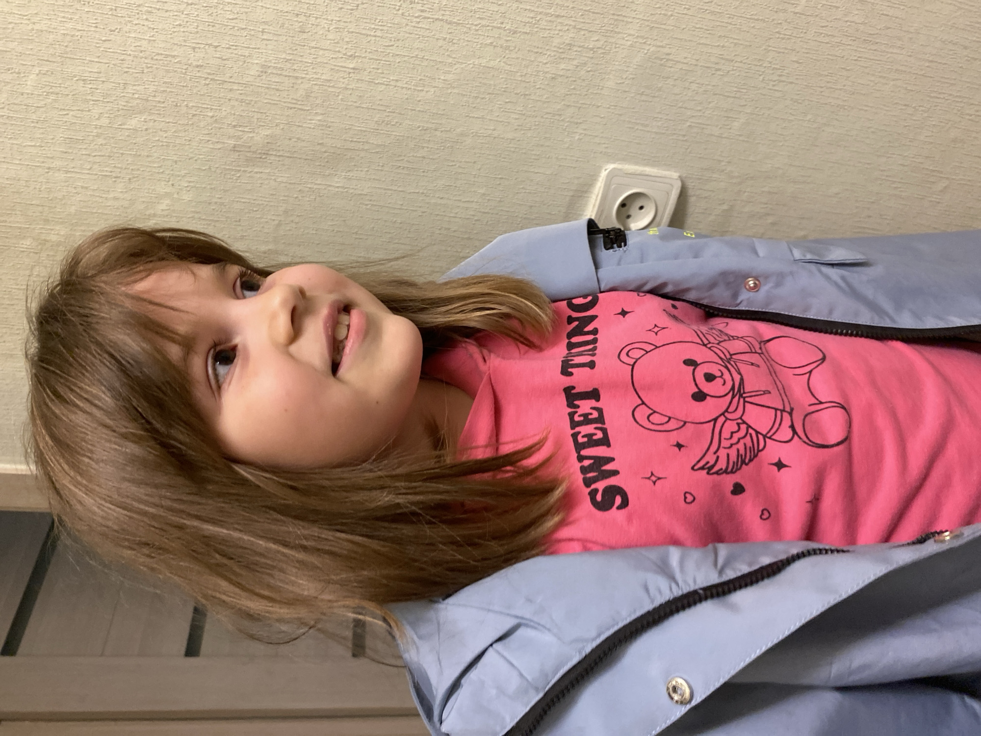 A medium shot depicts a young girl indoors. She is smiling slightly, her gaze directed upwards. The girl is wearing a pink t-shirt adorned with a teddy bear design, and a blue jacket. The background features a light-colored wall with a white electrical outlet. The image's orientation is sideways. The location is not specified within the image; it is an unsupported request.