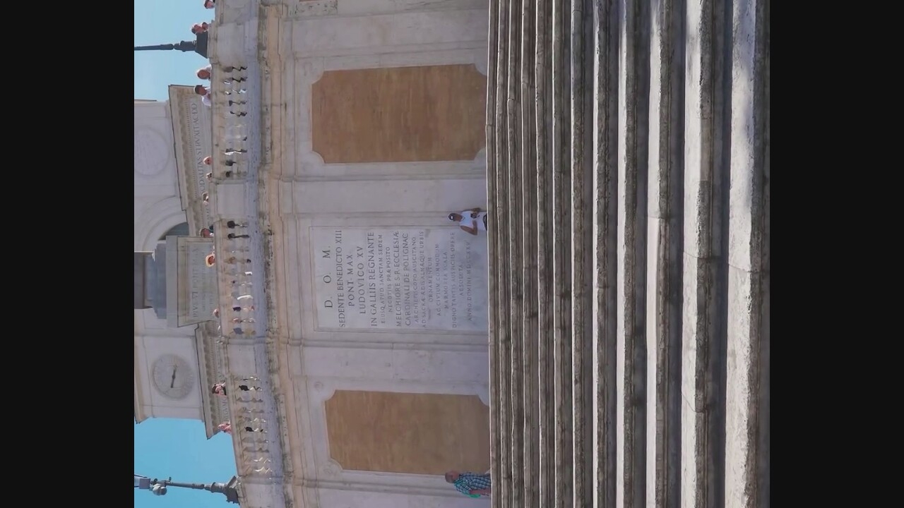 This is an eye-level, medium shot of the Spanish Steps in Rome, Italy, on a sunny day. The steps, made of stone, take up the majority of the foreground and extend upwards towards a building with a large inscription. The inscription is in Latin and appears to be a dedication. Above the inscription, a balustrade runs along the building, with several people standing on it, looking out. One person is seated on the steps, looking at their phone. Another person, wearing a blue and white checkered shirt, walks up the steps. The sky is clear blue.