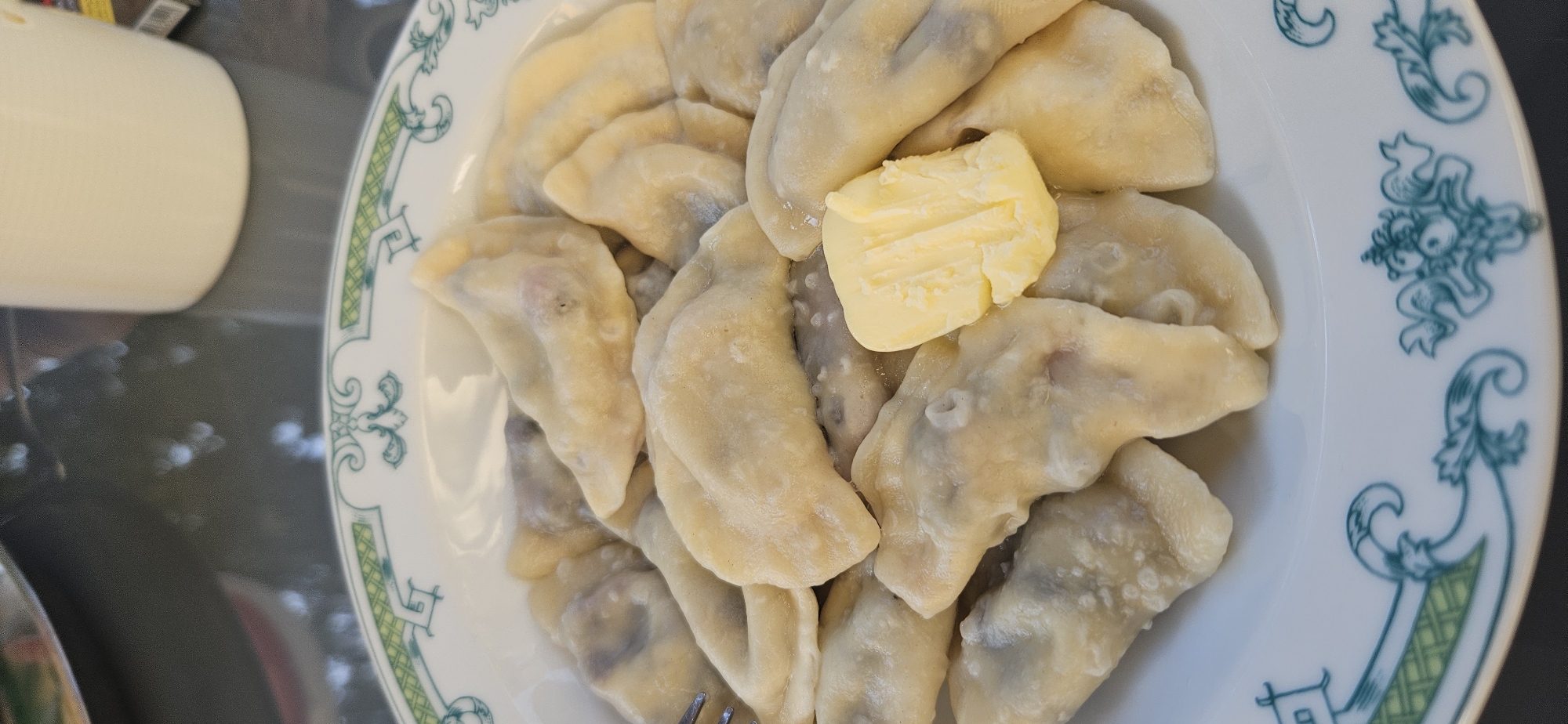 A plate of dumplings sits on a table in Hévíz, Hungary. The dumplings appear to be filled with a dark substance, possibly meat or fruit, and are topped with a pat of yellow butter. The plate has a decorative green and white rim. In the background, a white mug and a portion of another plate are visible. The table surface is dark and reflective, showing blurred shapes of light. The lighting suggests an indoor setting, possibly a restaurant or a home.