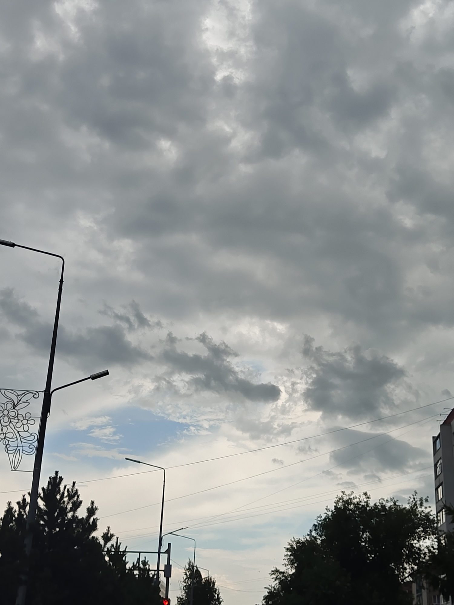 The image depicts an overcast sky with a building on the right and trees in the foreground. A lamppost with two lights is visible on the left, and another lamppost is in the center. The sky is filled with dense, gray clouds, with a small patch of blue visible near the horizon. The building appears to be a multi-story structure, possibly an apartment building, with windows reflecting the dim light. The trees are dark green and appear to be coniferous. The scene suggests a cloudy day in an urban environment. The location in the city of Petropavl, Kazakhstan, cannot be determined from the visual information provided in the image.