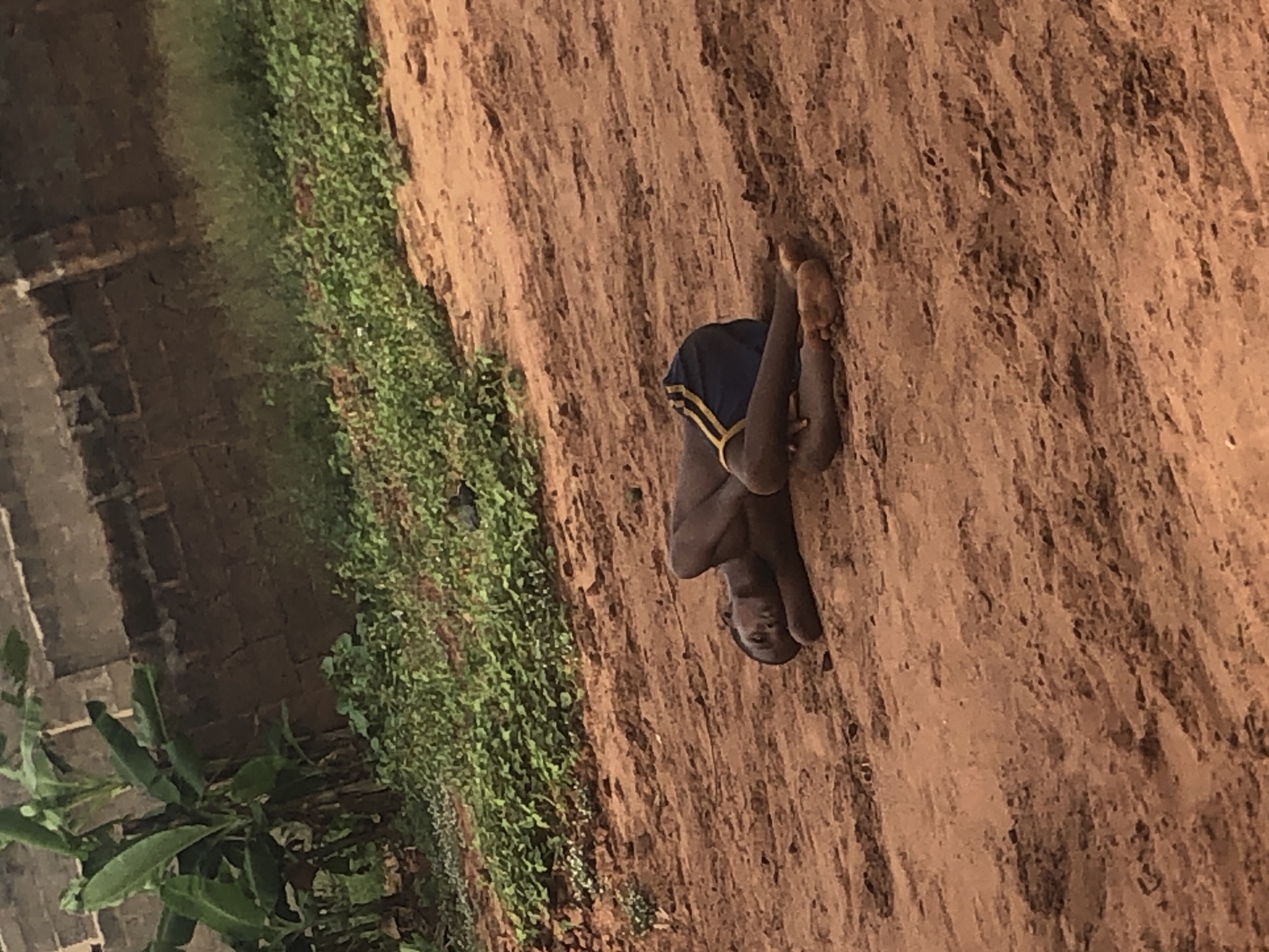 The image, taken in Ukwu-Abwa, Nigeria, portrays a young person, likely a boy, sitting on reddish-brown dirt ground. The individual has dark skin and is wearing blue shorts with yellow striped details. He is positioned with his knees drawn up towards his body, and he looks directly at the camera with a neutral expression.
The setting is an outdoor, unpaved area. The ground is textured earth, suggesting a natural and possibly rural or semi-urban environment. To the left side of the frame, there is a patch of green vegetation, composed of small plants and leaves, bordering the dirt area. Further left, a dark-colored brick or block structure, possibly a wall or part of a building, is visible. In the bottom-left corner, several large green leaves, characteristic of a banana plant, are present.
The scene is illuminated by natural daylight, indicating it is daytime. The weather appears fair. There are no other discernible activities or individuals. No text is visible in the image.