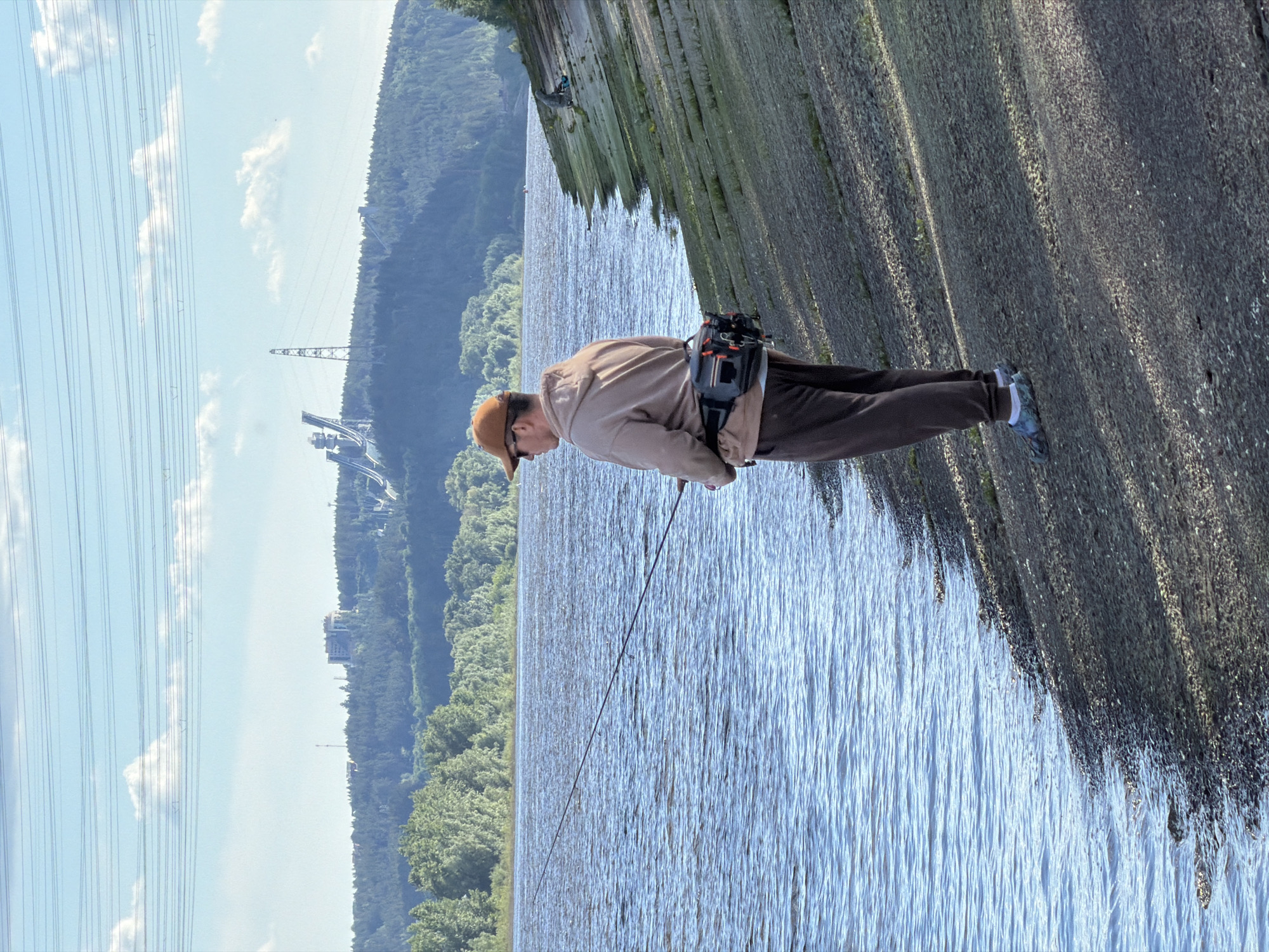 A man fishes on the embankment of the Kama River in the city of Chaikovsky, Russia. He wears a brown cap, tan jacket, and brown pants. A fishing pole extends into the water. In the background, transmission towers are visible against a partly cloudy sky. The river is lined with trees. The embankment is concrete.