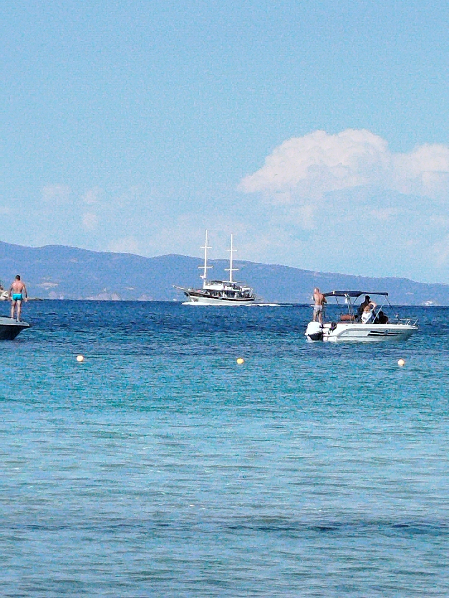 The image depicts a serene coastal scene in Sithonia Municipal Unit, Greece. The foreground is dominated by the tranquil, turquoise waters of the sea, with subtle ripples suggesting a gentle breeze. In the middle ground, two boats are visible. To the left, a man in blue shorts stands on the edge of a dark boat, appearing to be either preparing to dive or simply enjoying the view. Further to the right, a white motorboat with a canopy carries several people, engaged in conversation or relaxation. In the background, a larger, traditional sailing vessel with multiple masts cruises across the deep blue water. Beyond the sea, a range of hazy, blue-green mountains forms the horizon under a clear sky dotted with a few wispy white clouds. The overall impression is one of a leisurely summer day at sea.