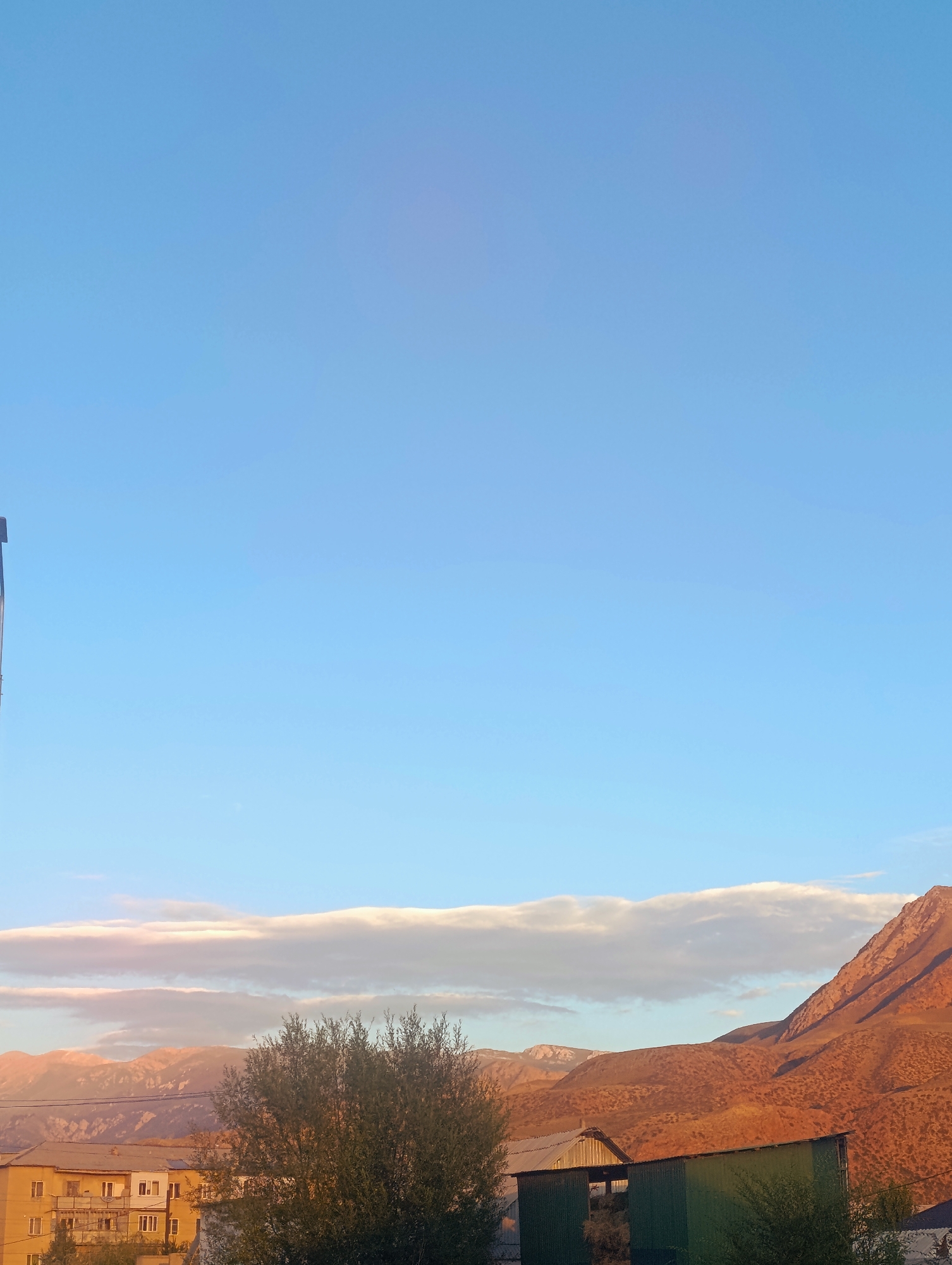 The image captures a serene daytime scene in Naryn City, Kyrgyzstan, with a clear, pale blue sky dominating the upper half. The lower portion of the image reveals a landscape characterized by rolling hills and distant mountains bathed in the warm, golden light of the sun, suggesting it might be late afternoon or early morning.
In the foreground, a multi-story apartment building with a pale facade and numerous windows stands to the left. Adjacent to it, a lush, green tree provides a touch of natural vibrancy. Further to the right, two simple, barn-like structures with corrugated metal roofs are visible, one appearing to be open-sided, possibly used for storage. The overall environment suggests a semi-rural or suburban setting within the city, with buildings interspersed with natural elements. The absence of people or specific actions indicates a static, observational view of the locale.