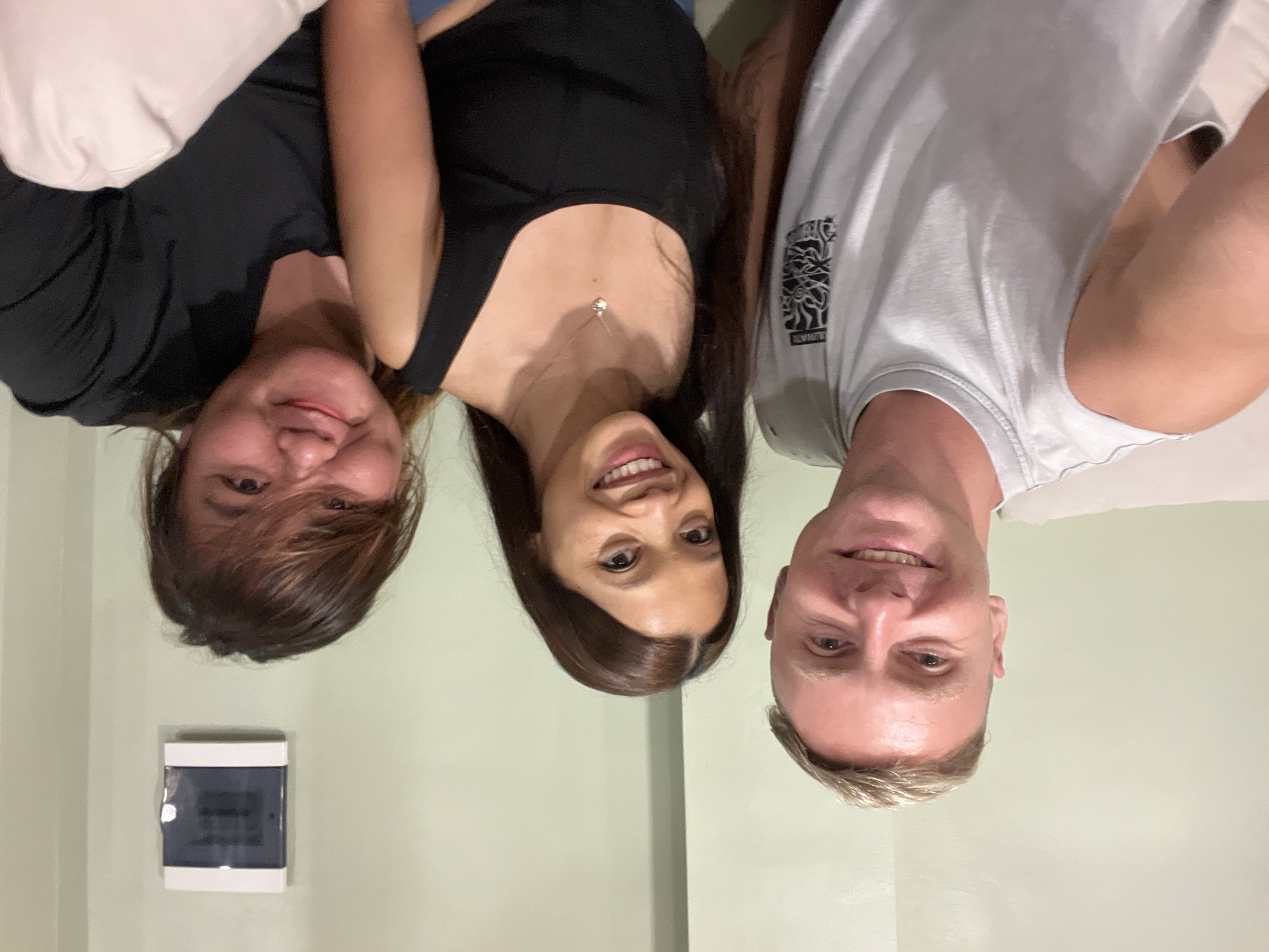 Three people are posing closely together. The person on the left has brown hair and is wearing a black top. The person in the middle has long dark hair and is wearing a black tank top with a silver necklace. The person on the right has short blonde hair and is wearing a white t-shirt. A white object is visible in the background, along with light green walls. The photo was taken indoors, and the image is rotated sideways. The location is potentially North Kuta, Indonesia.