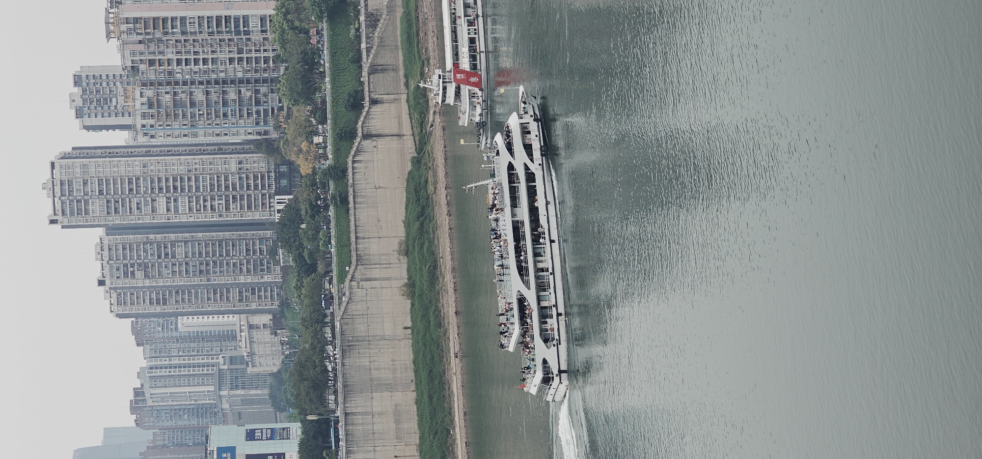 The scene depicts a riverfront cityscape in Yuzhong District, China. A large passenger ship, possibly a cruise boat, is moving along the river, with numerous people visible on its decks. The river is wide and calm, reflecting the overcast sky. On the left bank, a concrete embankment runs alongside a narrow strip of greenery with trees. Behind this, a dense cluster of tall apartment buildings dominates the skyline. The weather appears overcast and hazy.