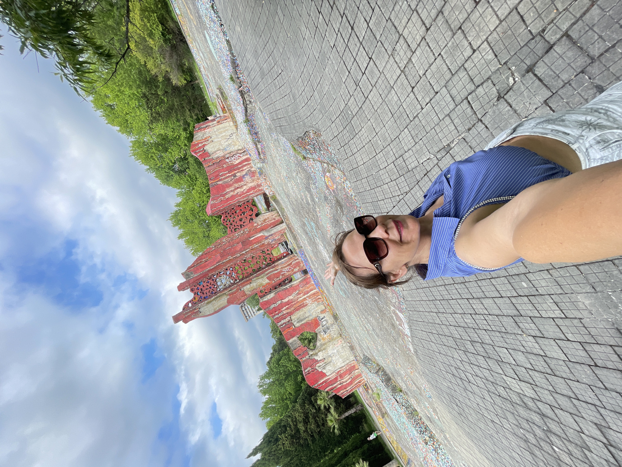 An outdoor, eye-level, wide-angle selfie shows a woman in her 40s wearing a blue striped shirt, white pants, and sunglasses. She is smiling and holding the camera in her right hand, while her left hand is raised with her index finger extended, pointing towards a red and grey abstract sculpture in the background. The sculpture is partially covered in colorful mosaics and is situated in a public space with a paved ground made of grey interlocking tiles. Trees and a cloudy sky are visible behind the sculpture.