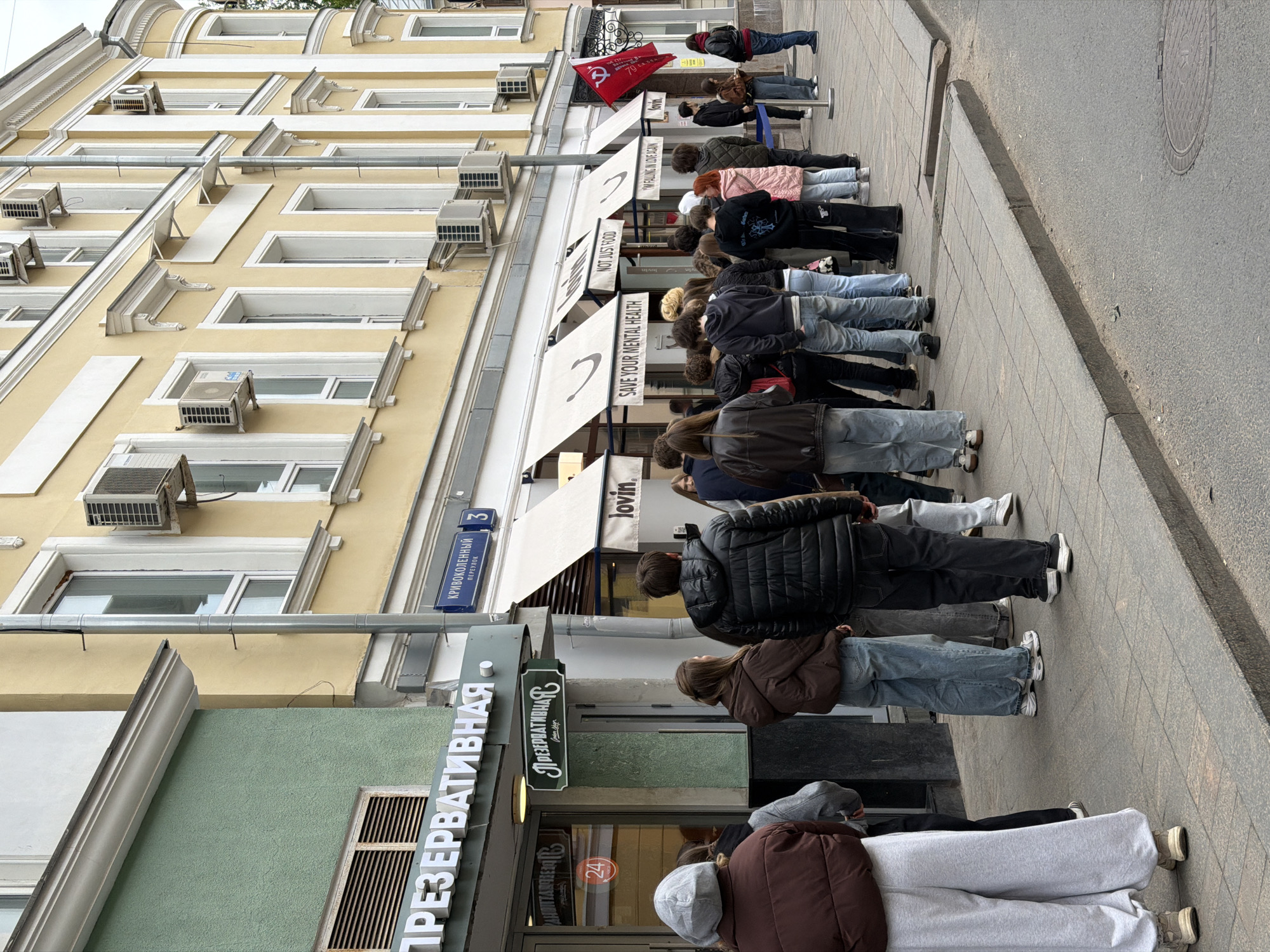 A queue of people stands on a Moscow, Russia, sidewalk in front of a shop identified by signage in Cyrillic and English. The first shop has the word "Презервативная" (PRESERVATIVNAYA) lit and visible. A red flag with Soviet symbols is visible near the front of the queue. The shop above has a sign with the word "lovin" on it. The awnings of the second shop have phrases such as "SAVE YOUR MENTAL HEALTH" and "NOT JUST FOOD" in English. The building is tan with white framed windows and air conditioning units.