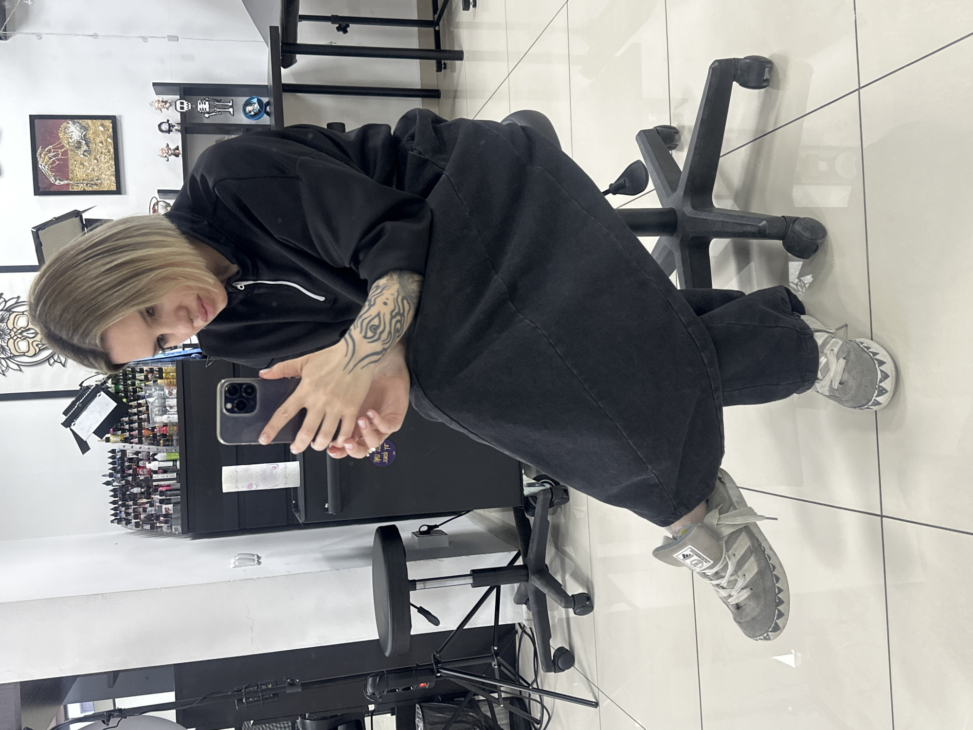 A young adult female with short, blonde hair is depicted seated on a black, wheeled adjustable stool within what appears to be a tattoo studio located in Rostov-on-Don, Russia. She is wearing a black hooded sweatshirt with a white zipper, black loose-fitting pants, and grey Adidas sneakers featuring a distinctive white, jagged shark-tooth pattern on the toe caps. Visible tattoos adorn her left forearm. She holds a dark-colored smartphone, consistent with an iPhone Pro model, with both hands and is directing her gaze downwards towards its screen. The environment consists of white walls and a white tiled floor. In the background, a black cabinet displays numerous small bottles, likely tattoo inks, neatly arranged. Several framed artworks, including one depicting a skeletal hand, are mounted on the walls. Additional visible items include a roll of white paper towels, small figurines on a shelf, a black light stand or tripod, and a metal-framed table.