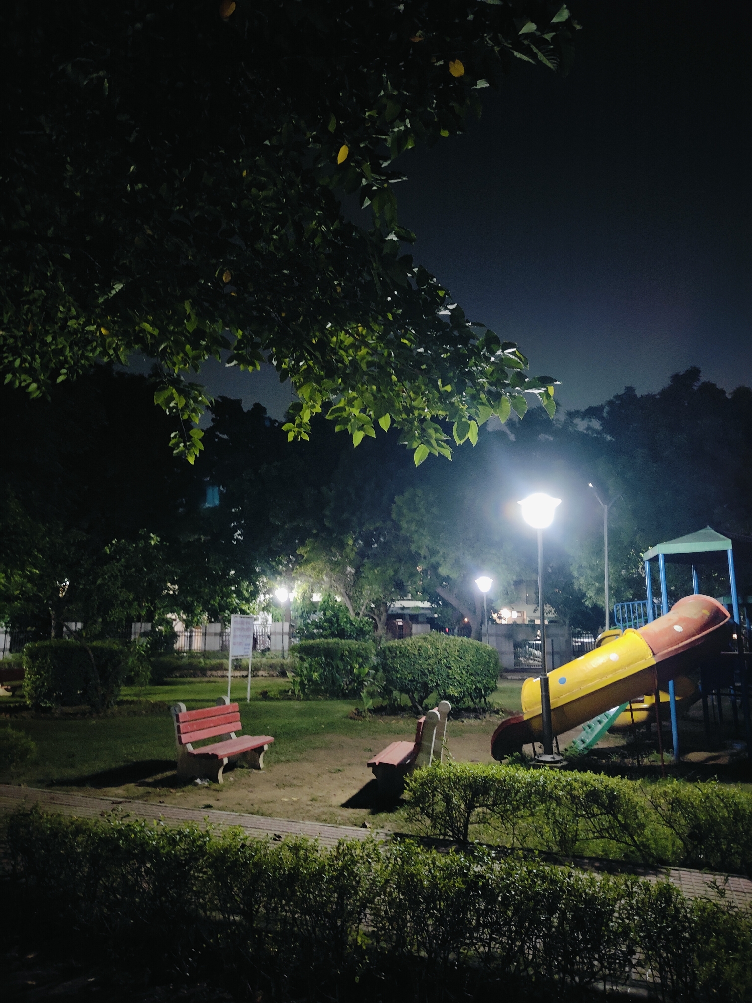 The image depicts a children's playground at night in Gurgaon, India. The scene is illuminated by streetlights and the glow of the night sky, creating a slightly eerie atmosphere. A yellow and red slide is the most prominent structure, with a blue support frame and green steps. To the left of the slide, there are two red benches, one with a backrest and the other without. Lush green bushes and trees surround the playground, their leaves dark against the night. The ground is a mixture of grass and dirt paths. In the background, a white fence and some buildings can be faintly seen through the trees. There are no people visible in the image, and no activity is taking place. The overall impression is one of an empty, quiet park after dark.