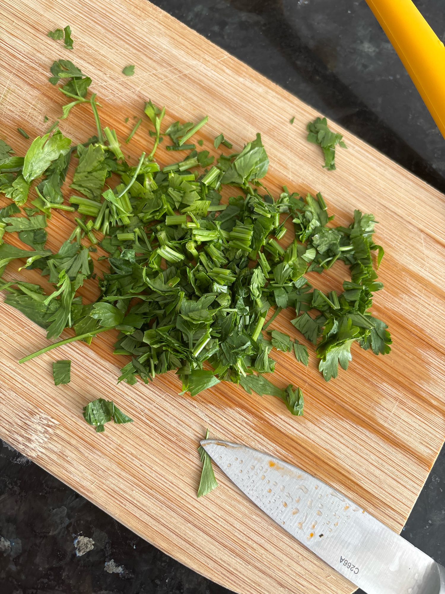 Chopped parsley is scattered across a wooden cutting board. A silver knife with water droplets and orange specks rests on the board. A yellow object, possibly a kitchen utensil, is visible in the upper right corner. The scene is set on a speckled dark countertop. The photograph was taken in the city of Málaga, Spain.