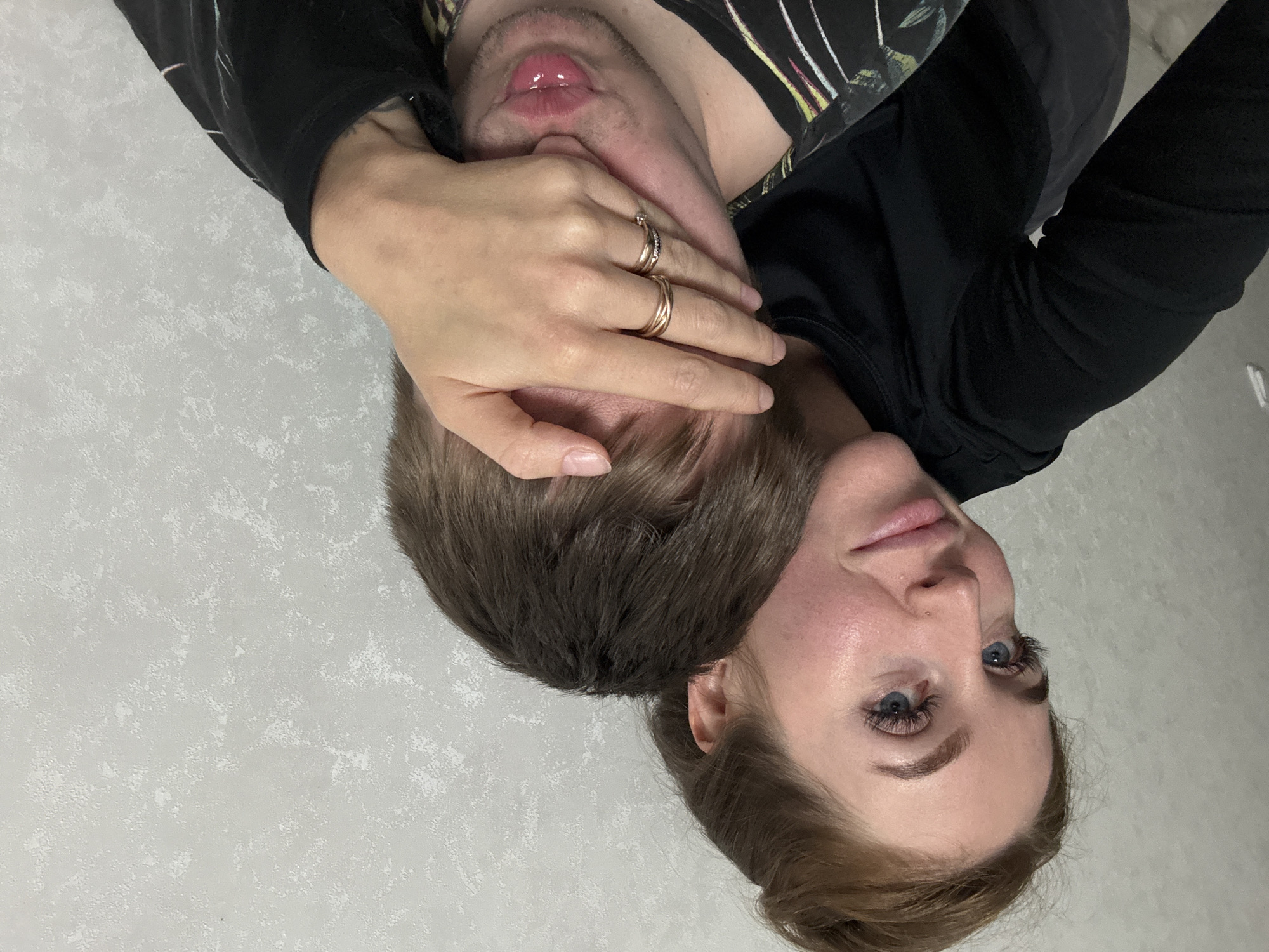 A close-up photo shows two people intertwined. A woman with fair skin, blue eyes, and reddish-brown hair styled in a bun is facing the camera. Her head is tilted slightly. A man's lips and part of his neck are visible above her, with a hand covering his face. The hand has two rings on the fingers. Both individuals are wearing dark-colored clothing. The background is a light, textured wall. There is no information to determine the location in the city of Chaikovsky, Russia.