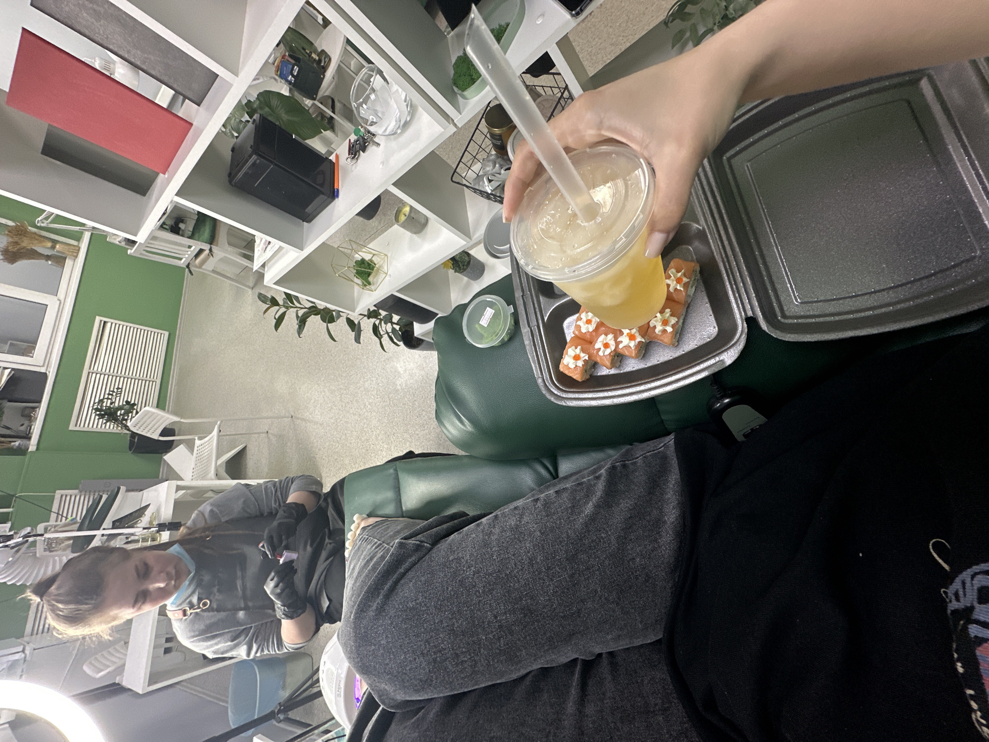 The image shows a person sitting in a green chair. They are wearing gray jeans and a black shirt. Their legs are crossed. To their right is a takeout container of sushi and a cup of a yellow drink with a straw. A hand is holding the drink. To the left of the person, in the background, is a person wearing gloves. They are wearing a gray shirt. The background appears to be a room with white shelves and some plants. The location is likely in Chaikovsky, Russia.
