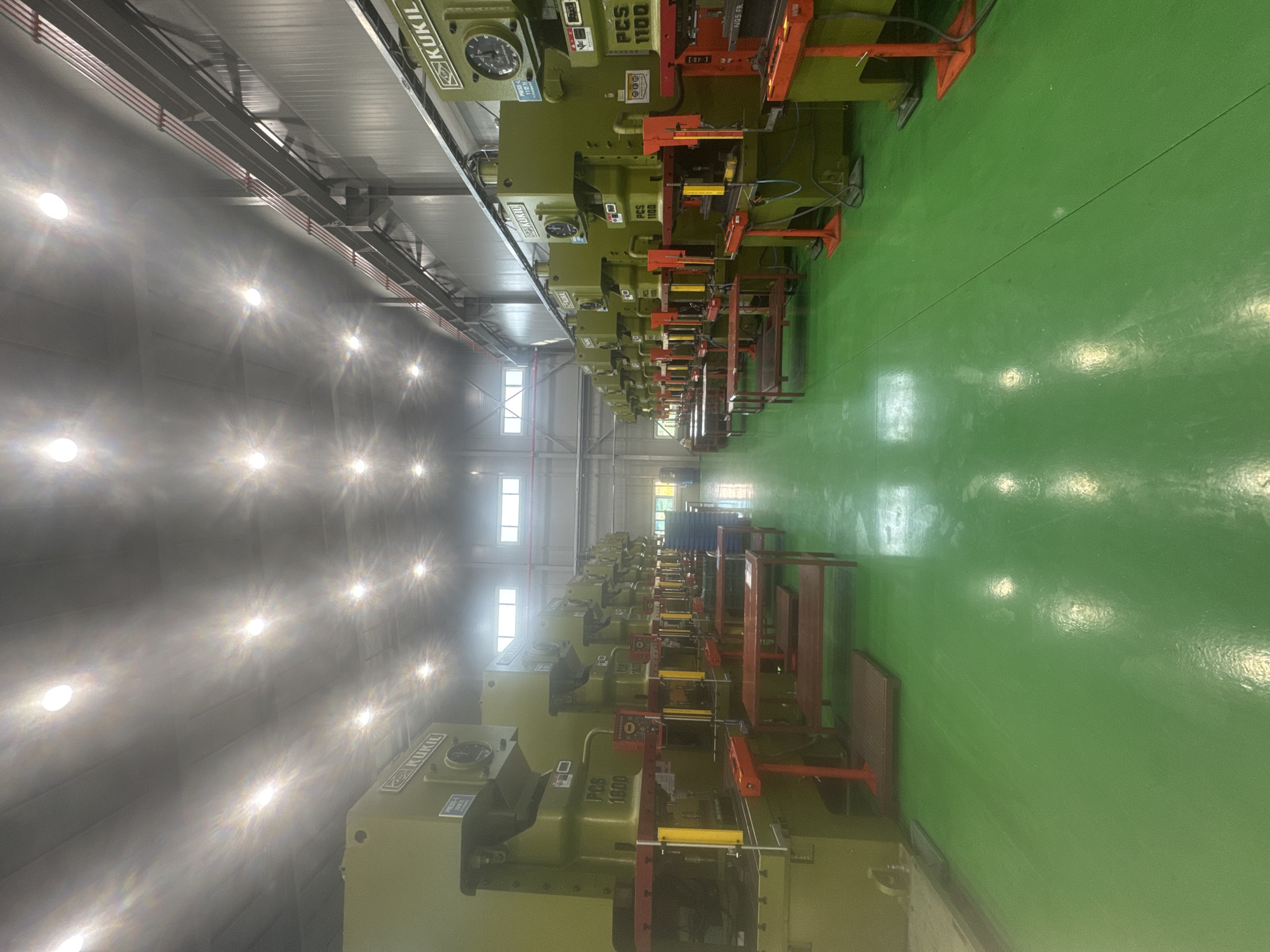 The image shows a row of industrial press machines in a factory setting. The machines are large, green, and appear to be of the "KUKIL" brand, with some labeled "PCS 1600" and "PCS 1100". They are arranged in a line, with their operational fronts facing the viewer. Red safety guards and metal shelving units are visible around some of the machines. The floor is a bright, glossy green. The ceiling is high with rows of circular fluorescent lights, some of which are creating glare. Windows are visible in the far wall. The overall environment is clean and organized, suggesting a manufacturing or production facility. The scene is located in the city of Pyeongtaek-si, South Korea, within a factory. There are no people visible in the image.