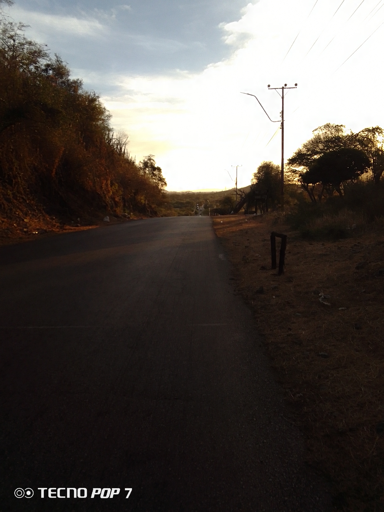 The image depicts a straight, asphalt road receding into the distance under a partially cloudy, sunlit sky. The road is flanked on the left by a hillside covered in dense vegetation. On the right, the road is bordered by dry grass and scattered trees. A utility pole with power lines is visible on the right side of the road. The photo was taken with a Tecno Pop 7 phone. The location is inferred to be near Altagracia, Venezuela, based on the prompt.