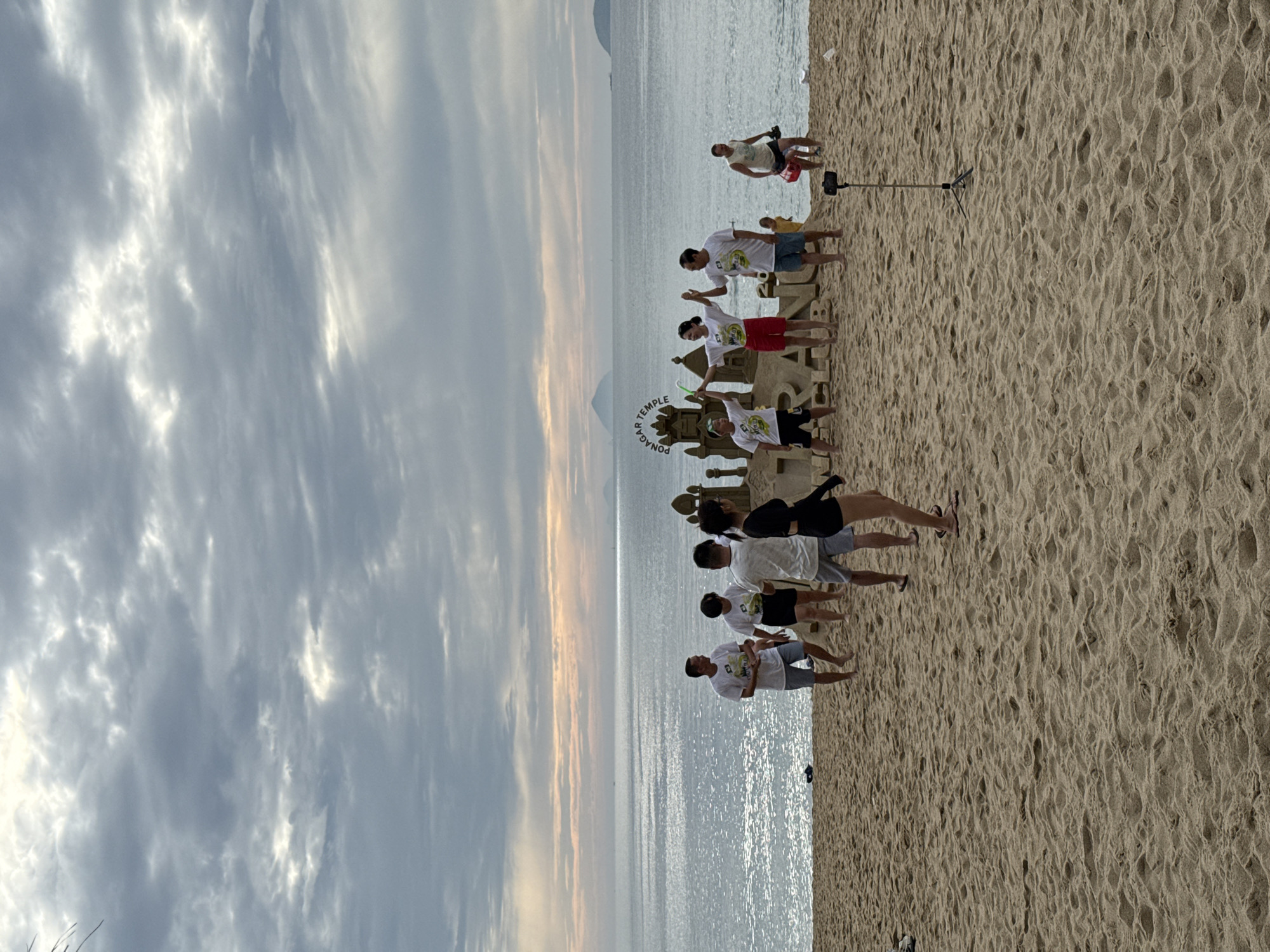 At the beach in Nha Trang, Vietnam, a group of people gather near a large sand sculpture of a temple that reads "PONAGAR TEMPLE" and "NHA TRANG CLUB". The sky is overcast with scattered sunlight breaking through. The ocean is visible in the background, with a few distant islands. Several people in the foreground are wearing white t-shirts, some with the same logo. Two women in the center are holding hands and appear to be posing. A man is walking in the background carrying a red bag. A tripod with a smartphone is set up on the sand in the foreground, suggesting the scene is being filmed or photographed. The sand is textured with visible footprints and ripples.