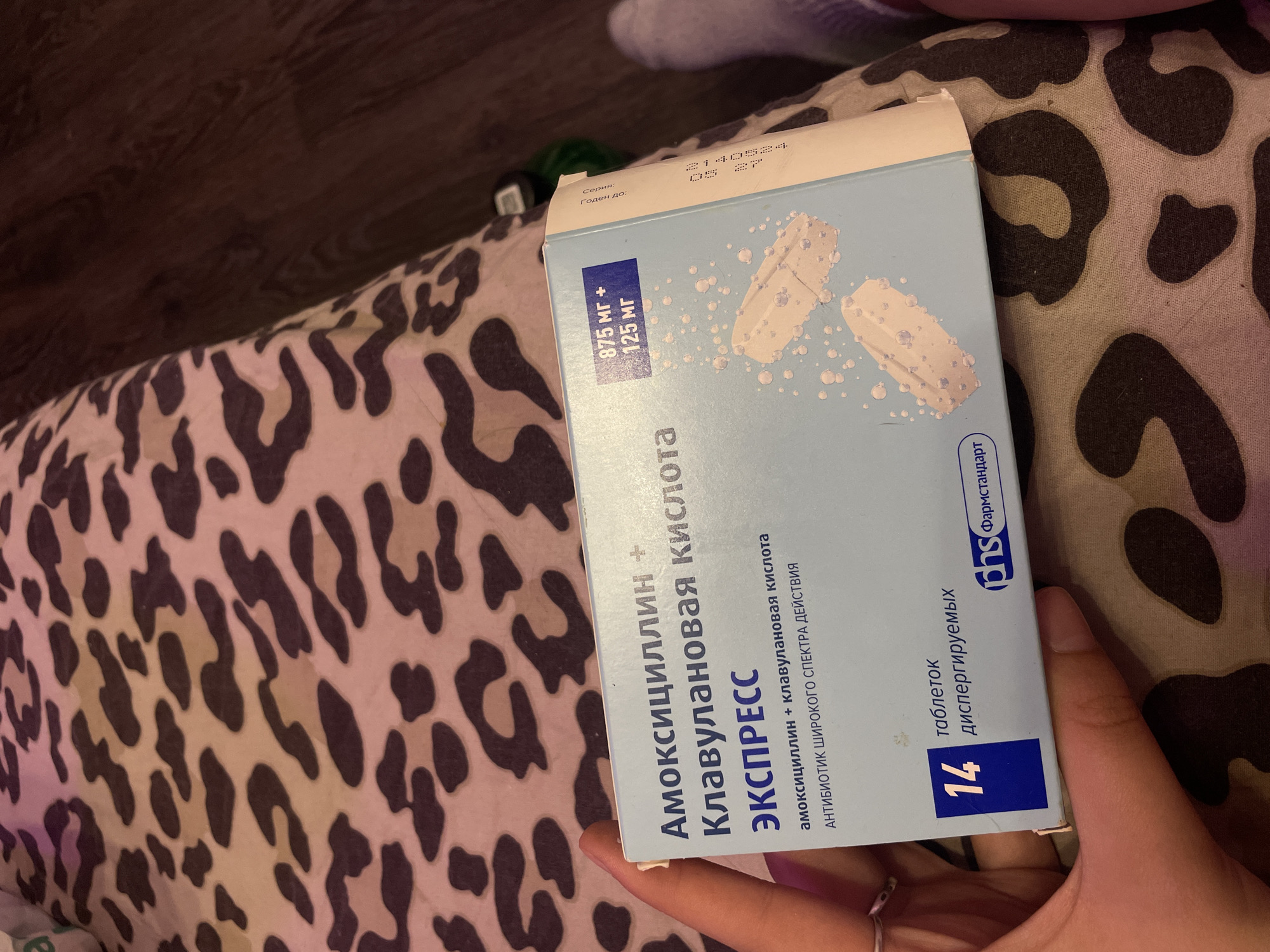 A box of Amoxicillin + Clavulanic Acid EXPRES is held by a person's left hand. The box is light blue with white lettering and depicts two white tablets with bubbles around them. The text on the box includes "Амоксициллин + Клавулановая кислота ЭКСПРЕСС", "875 мг + 125 мг", "14 таблеток", and "антибиотик широкого спектра действия". The box is placed on a leopard-print fabric. The background is dark and out of focus. The location is not specified in the image or provided context, so it cannot be determined to be in Vaganovo, Russia.